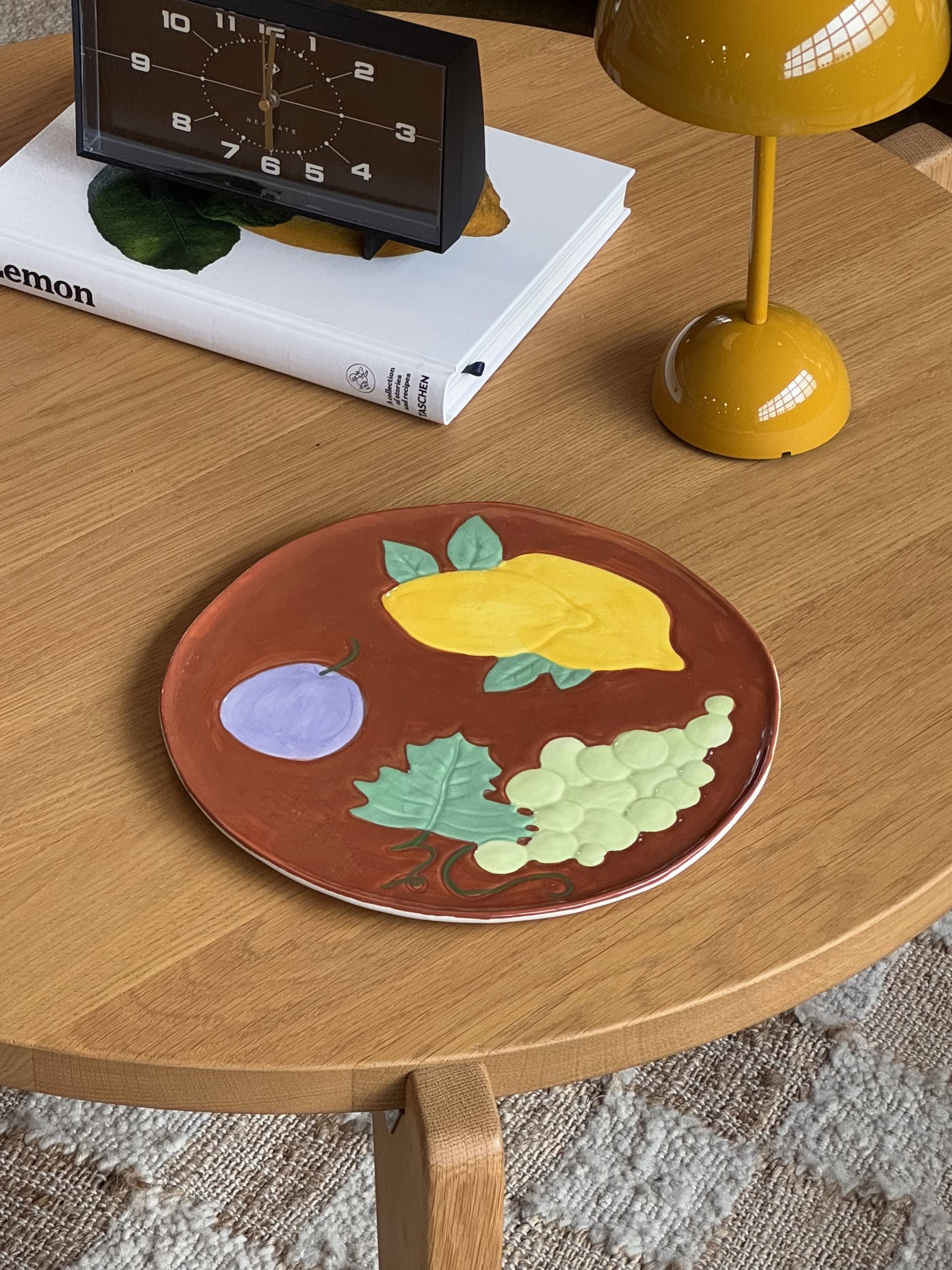 The &klevering Fruitful Grape Platter by &klevering, featuring a vintage fruit collage, adds vibrant colour to the wooden coffee table next to a yellow lamp, clock, and book.