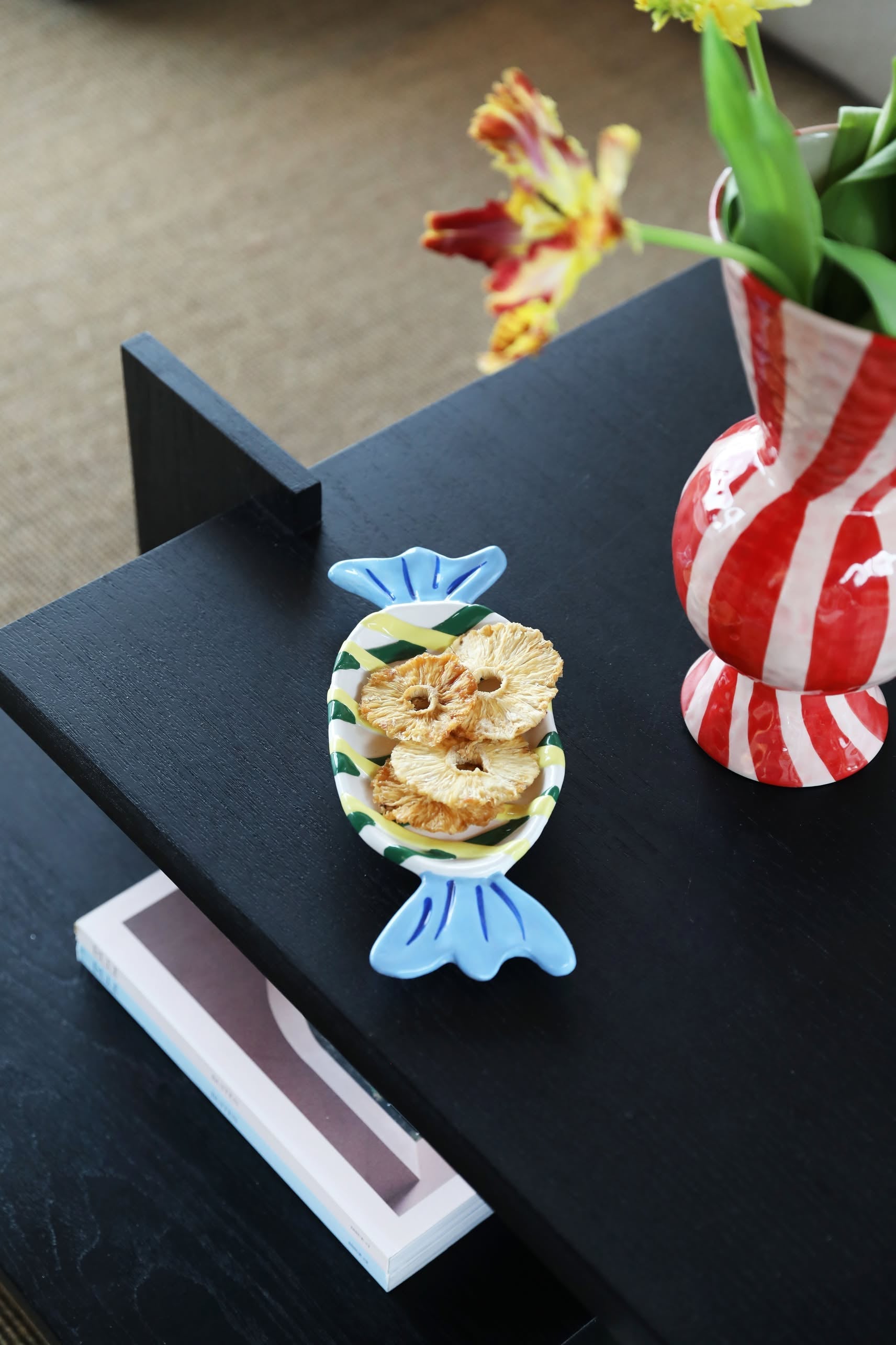 A medium &klevering Goody Plate with dried pineapple slices rests on a black table beside a red-and-white striped vase of yellow tulips, with a closed book on the shelf below for a whimsical touch.