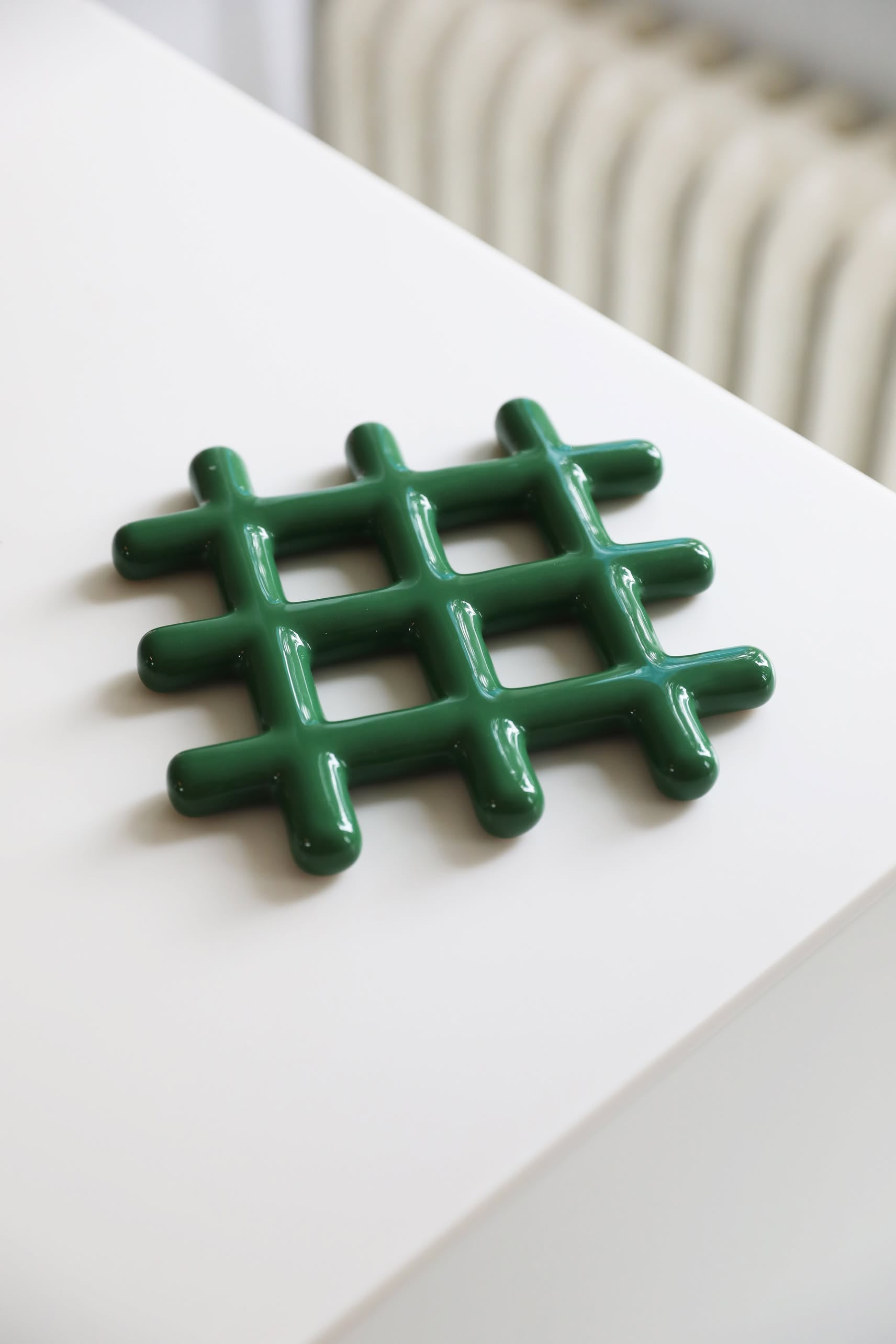 The &klevering Grid Trivet by &klevering, featuring a glossy dark green geometric design with rounded edges, sits on a white surface with a blurred radiator in the background.