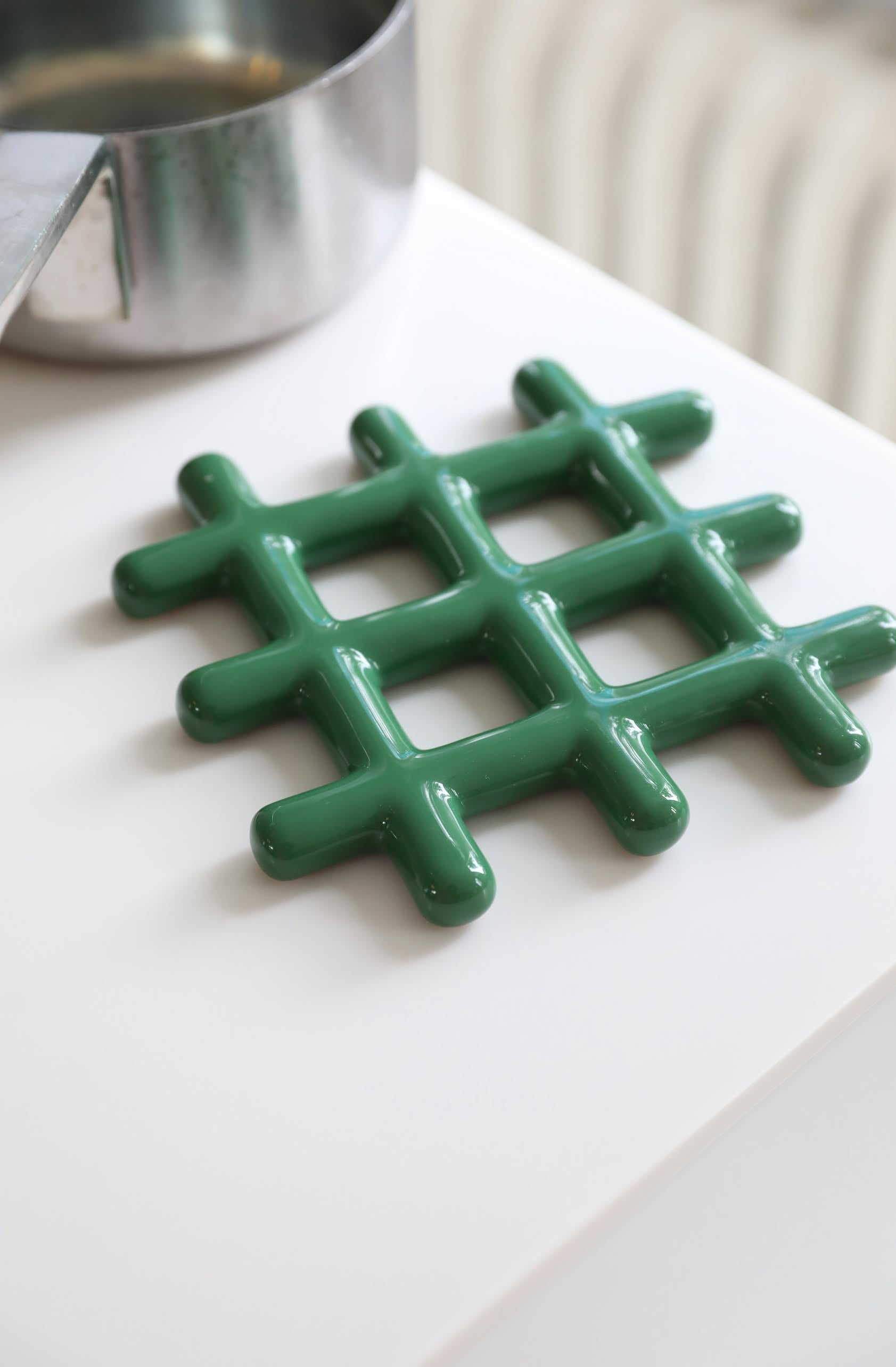 The &klevering Grid Trivet by &klevering, featuring a green glossy finish and geometric design, sits on a white surface next to a stainless steel pot, adding stylish heat protection.