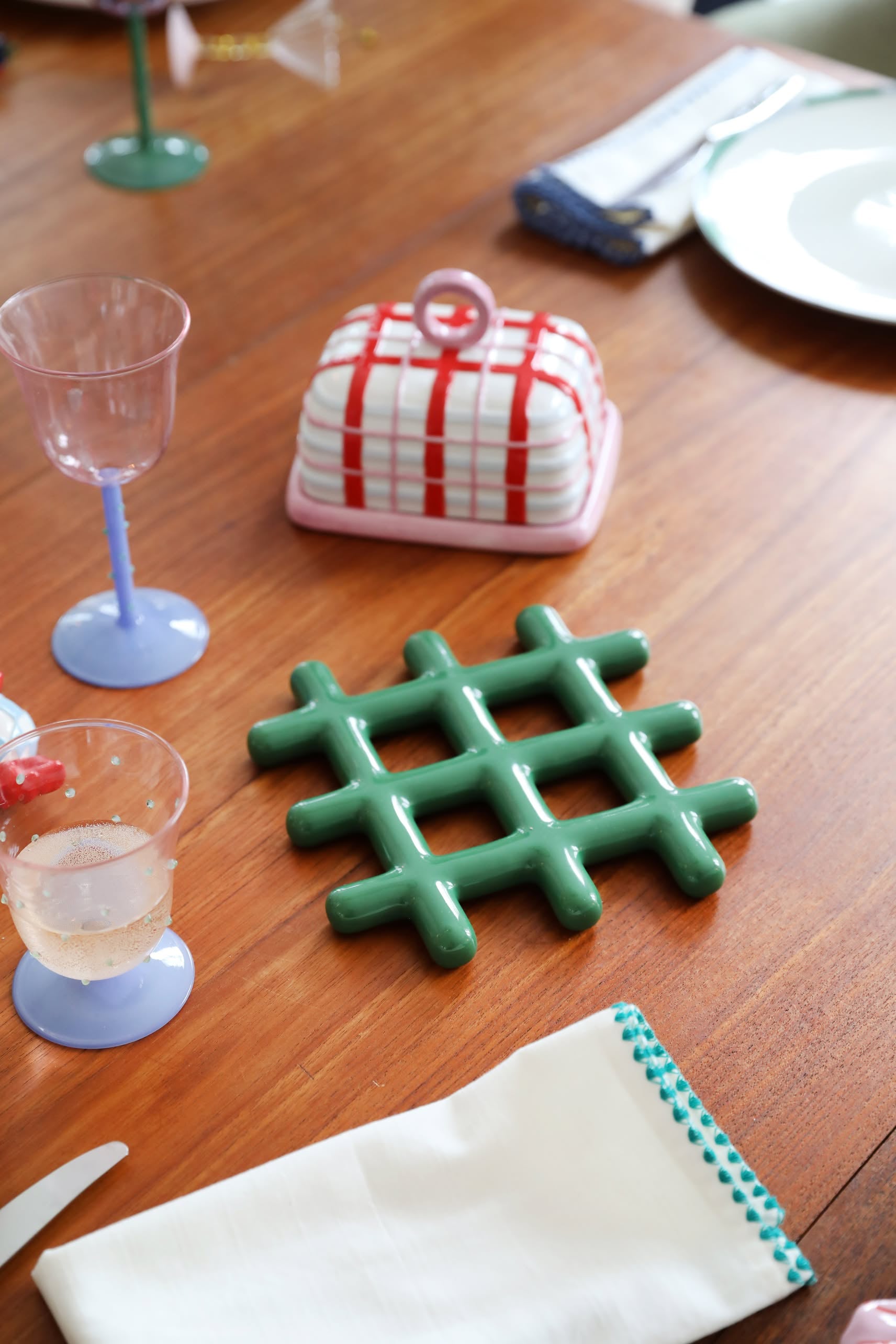 A wooden table set with pastel glassware, an &klevering Grid Trivet for heat protection with a geometric design, a checkered ceramic butter dish, a white napkin with blue stitching, and a partially visible plate.