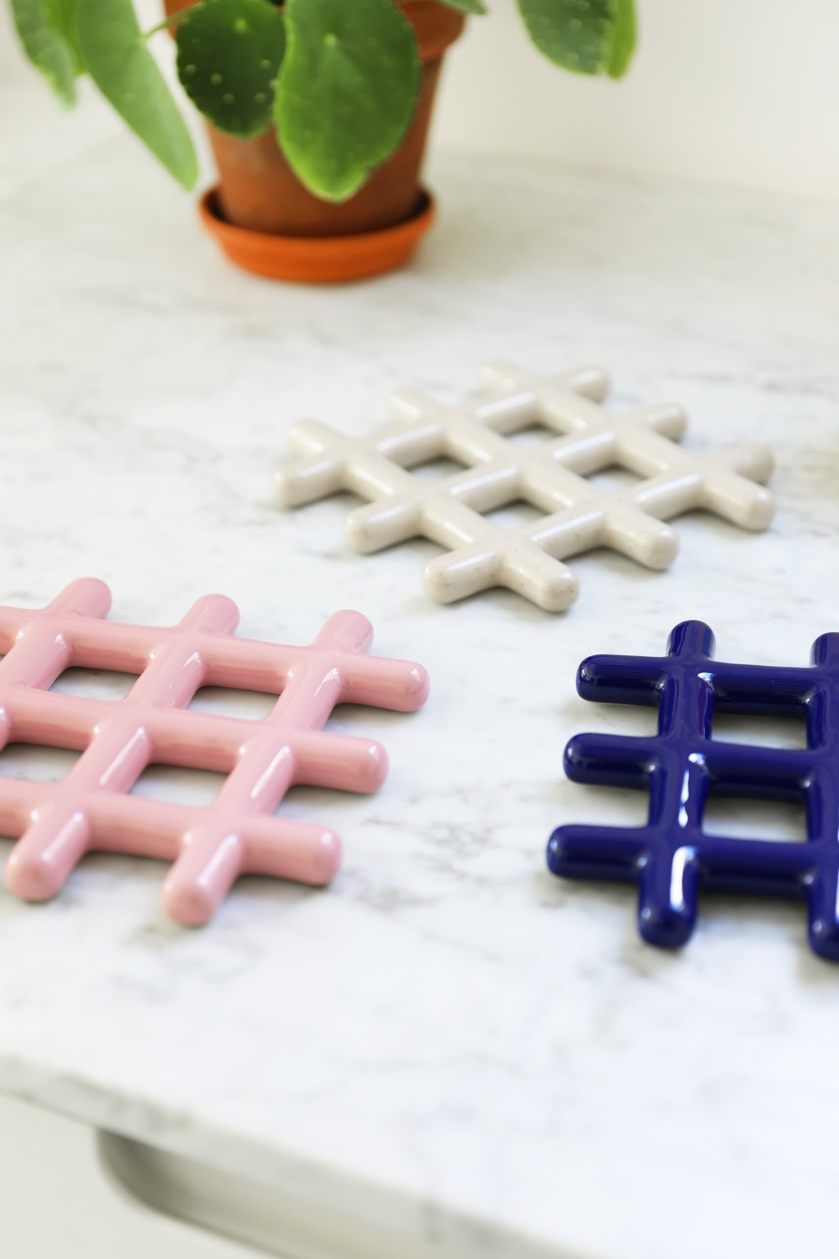 Three &klevering Grid Trivets in pink, white, and dark blue crisscross geometric designs by &klevering rest on a marble countertop, providing stylish heat protection. A potted plant sits in the background.