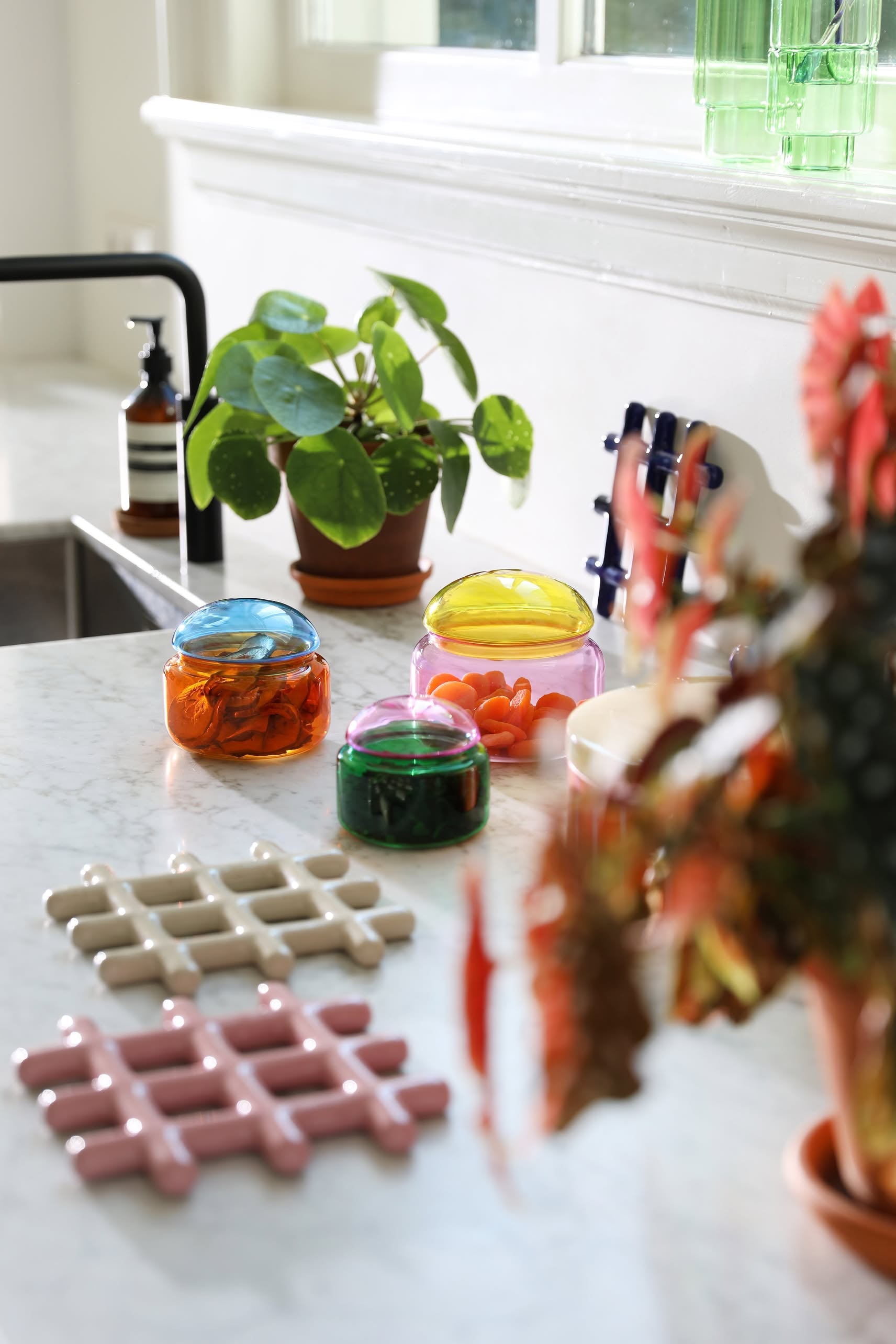 A bright kitchen counter displays plants in terracotta pots, colorful glass jars, and an &klevering Grid Trivet with a geometric design, while sunlight from a nearby window creates a warm, cozy feel.
