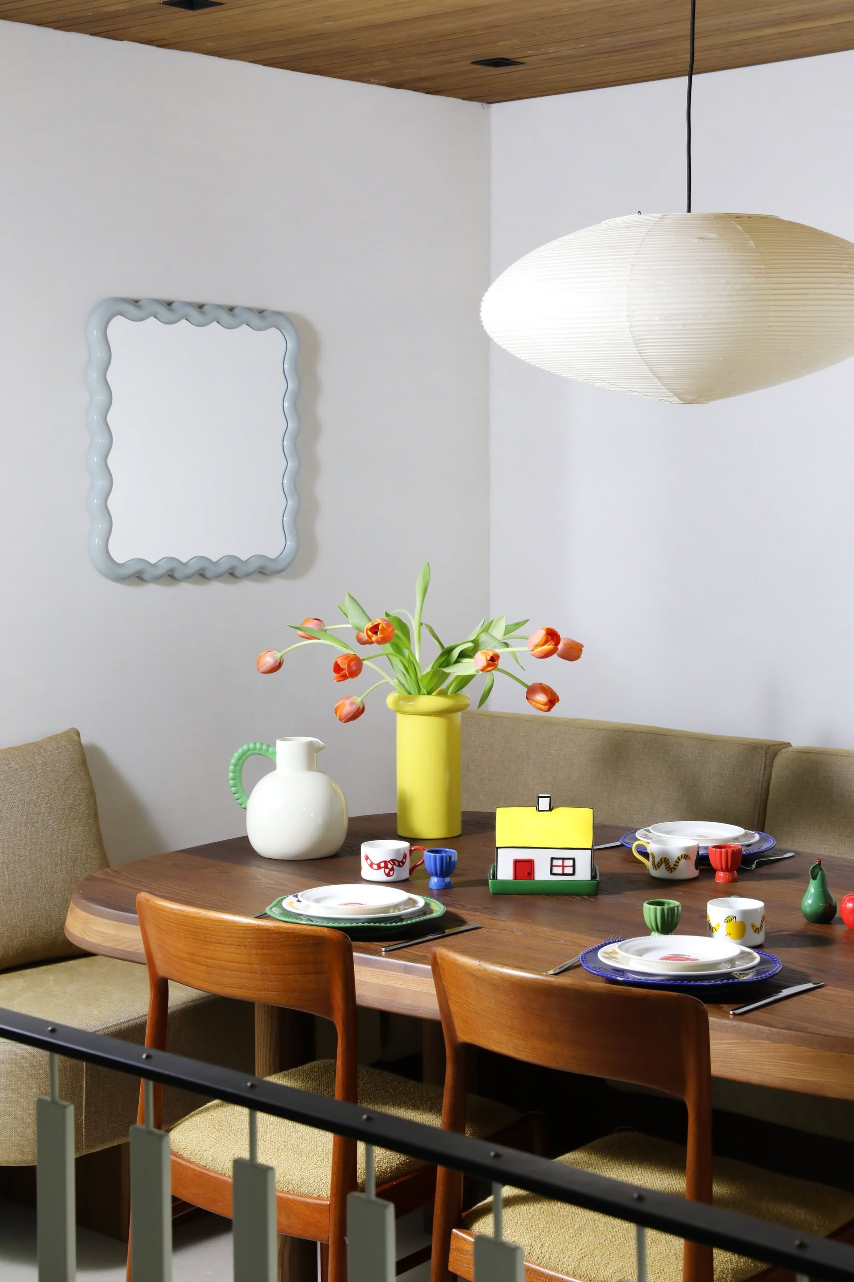 A modern dining room with a wooden English breakfast table for four, colorful dishes, an &klevering Haines Butter Dish, whimsical yellow vase with orange tulips, white jug, wavy wall mirror, cushioned seats, and large white pendant lamp overhead.