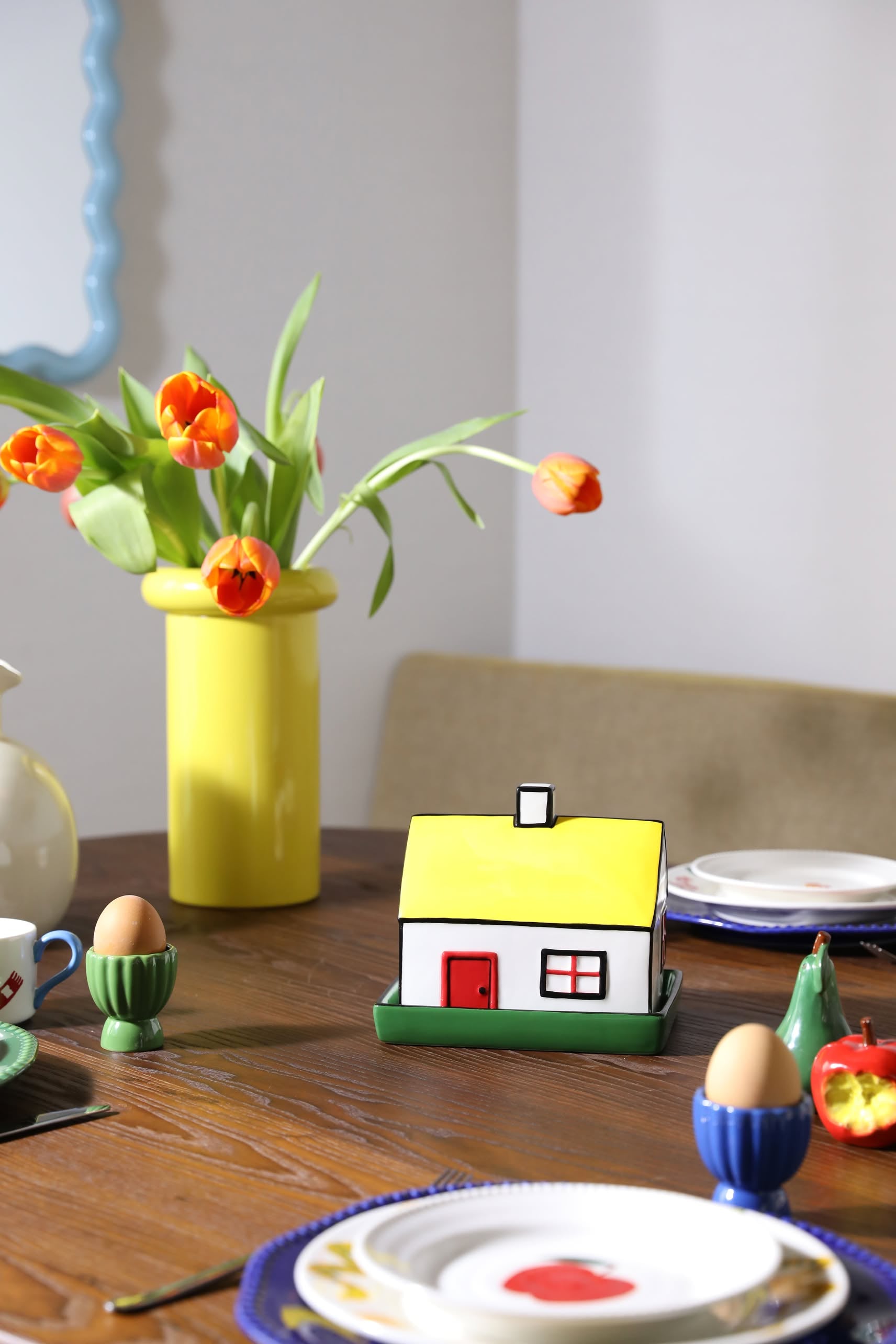 A wooden breakfast table is set with plates, egg cups, a vase of orange tulips, and the &klevering Haines Butter Dish by &klevering as a whimsical centerpiece. Sunlight streams in, brightening the inviting scene.