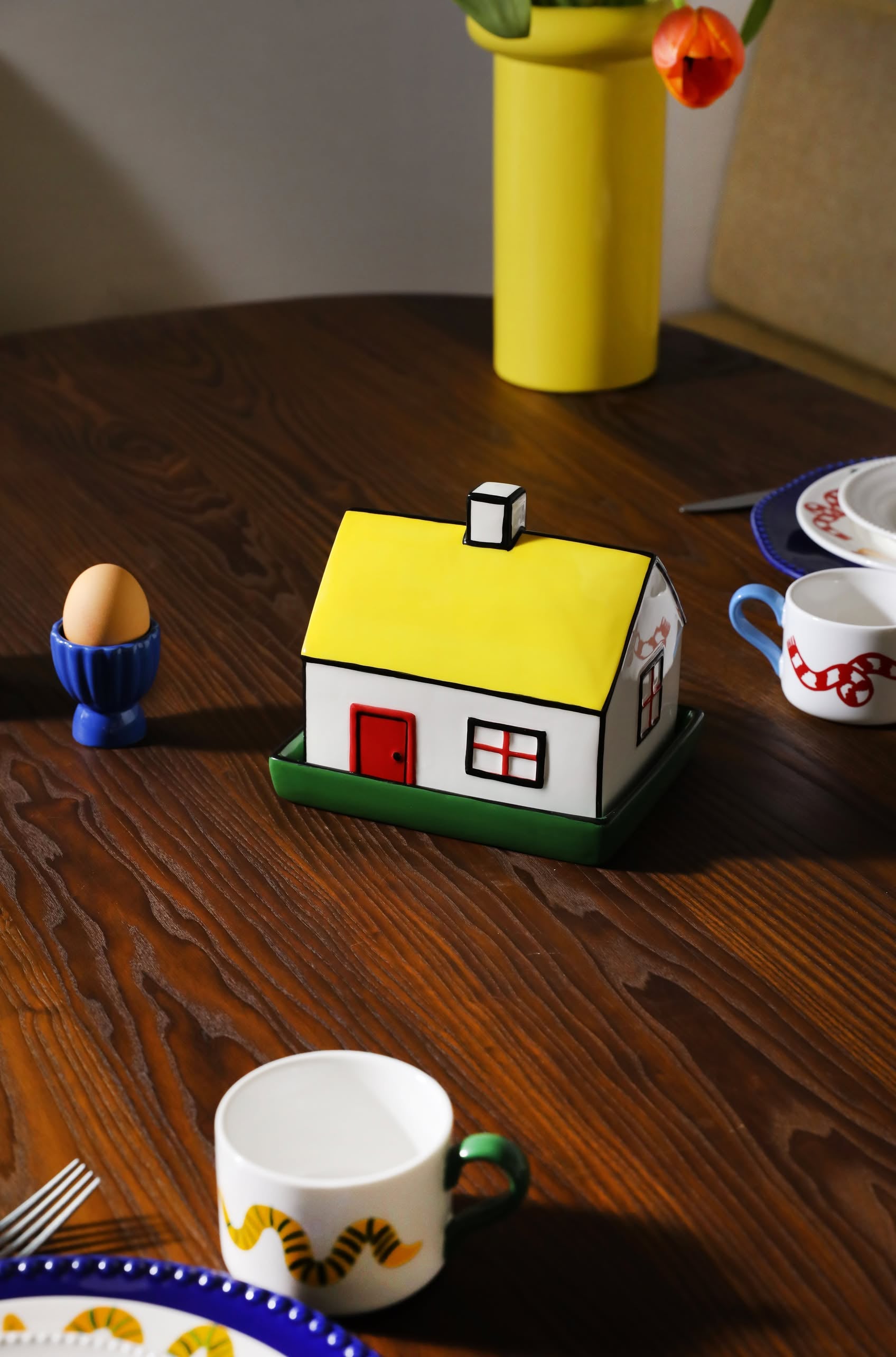 The &klevering Haines Butter Dish, with its house-inspired yellow roof design, rests on a wooden table surrounded by plates, a blue egg holder, and tulips—adding charm to any English breakfast setting.