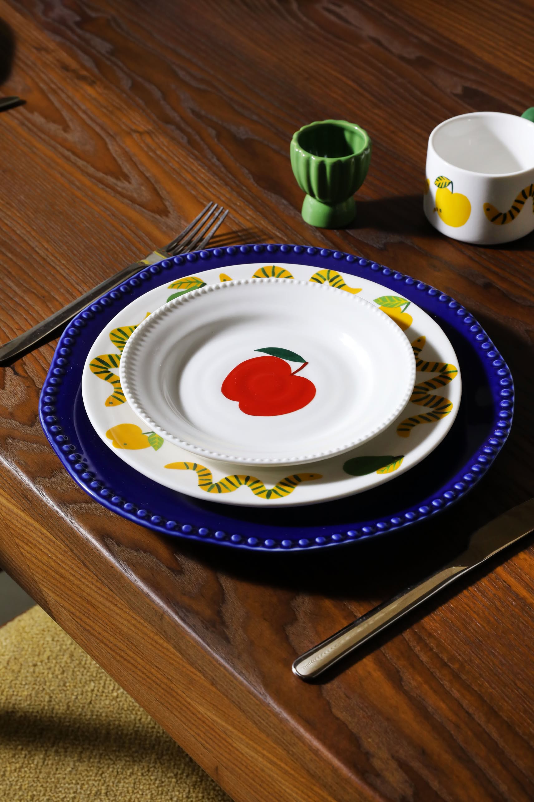 The &klevering Haines Plate Small, Set of 4 by &klevering features playful blue rims, yellow and red fruit patterns, and whimsical designs. Paired with cheerful tableware and cutlery, it creates a vibrant dining experience.