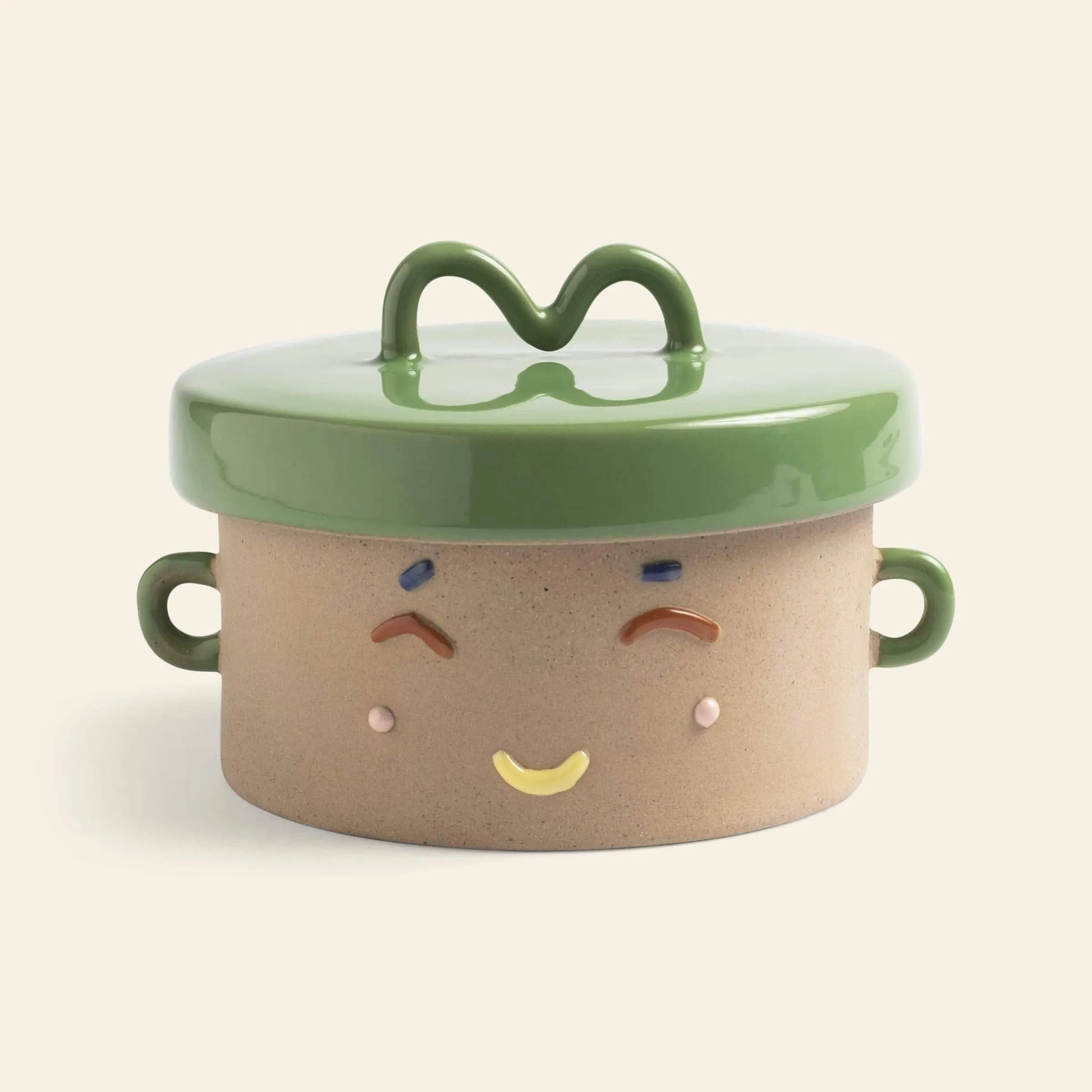 The &klevering Happy Jar by &klevering is a round ceramic jar with two handles, a green lid, and a smiling face with colorful painted details—a playful storage solution for any space.