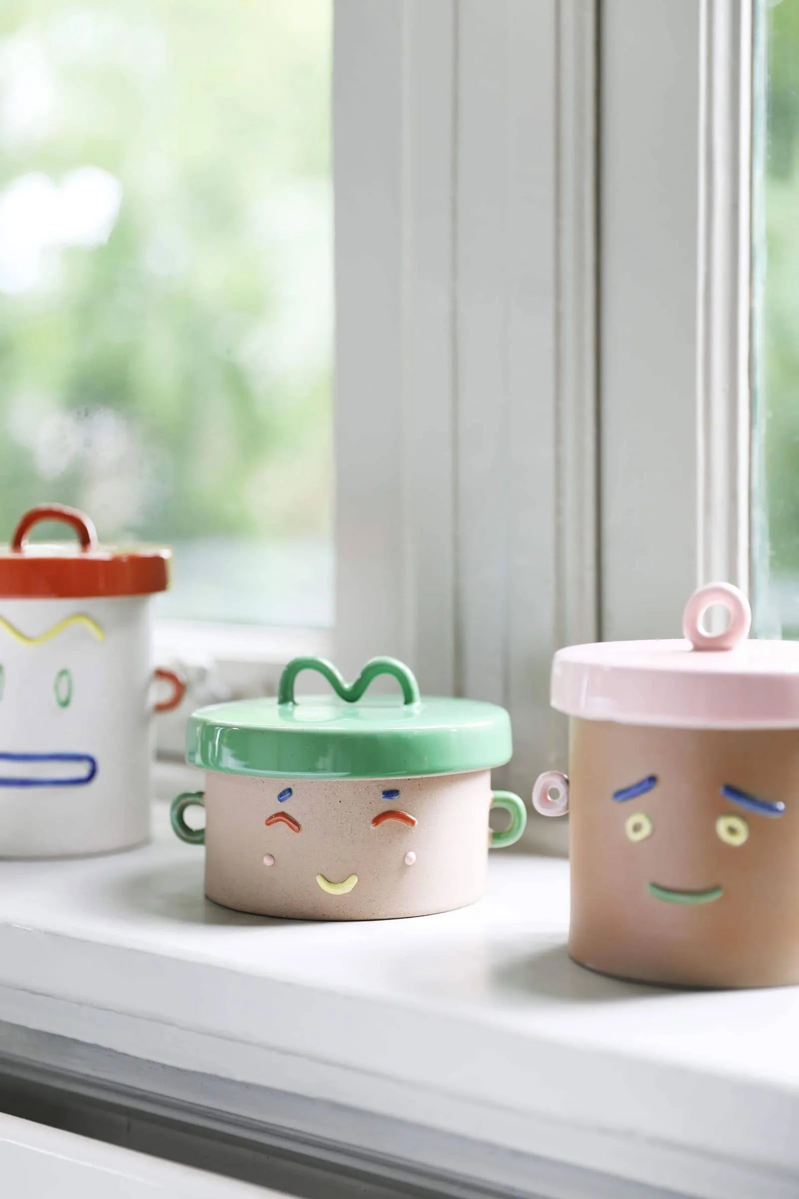 Three &klevering Happy Jars with colorful lids and handles sit on a windowsill. Each jar by &klevering has a unique facial expression, adding playful charm and versatile storage to your kitchen or living space.