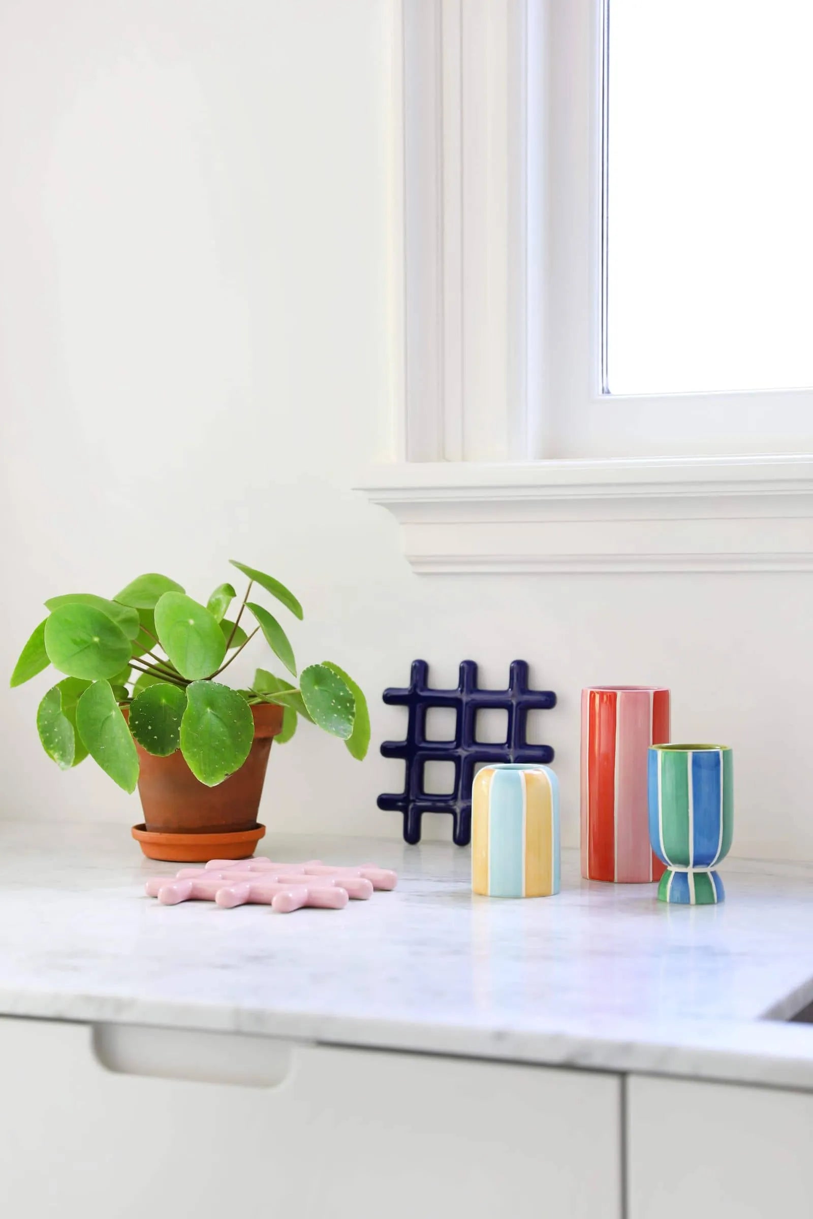 A potted plant and &klevering Ligne Vase, Set of 3 by &klevering are arranged on a white marble countertop near a window, along with a dark blue grid decor, pink trivet, and other geometric pieces.