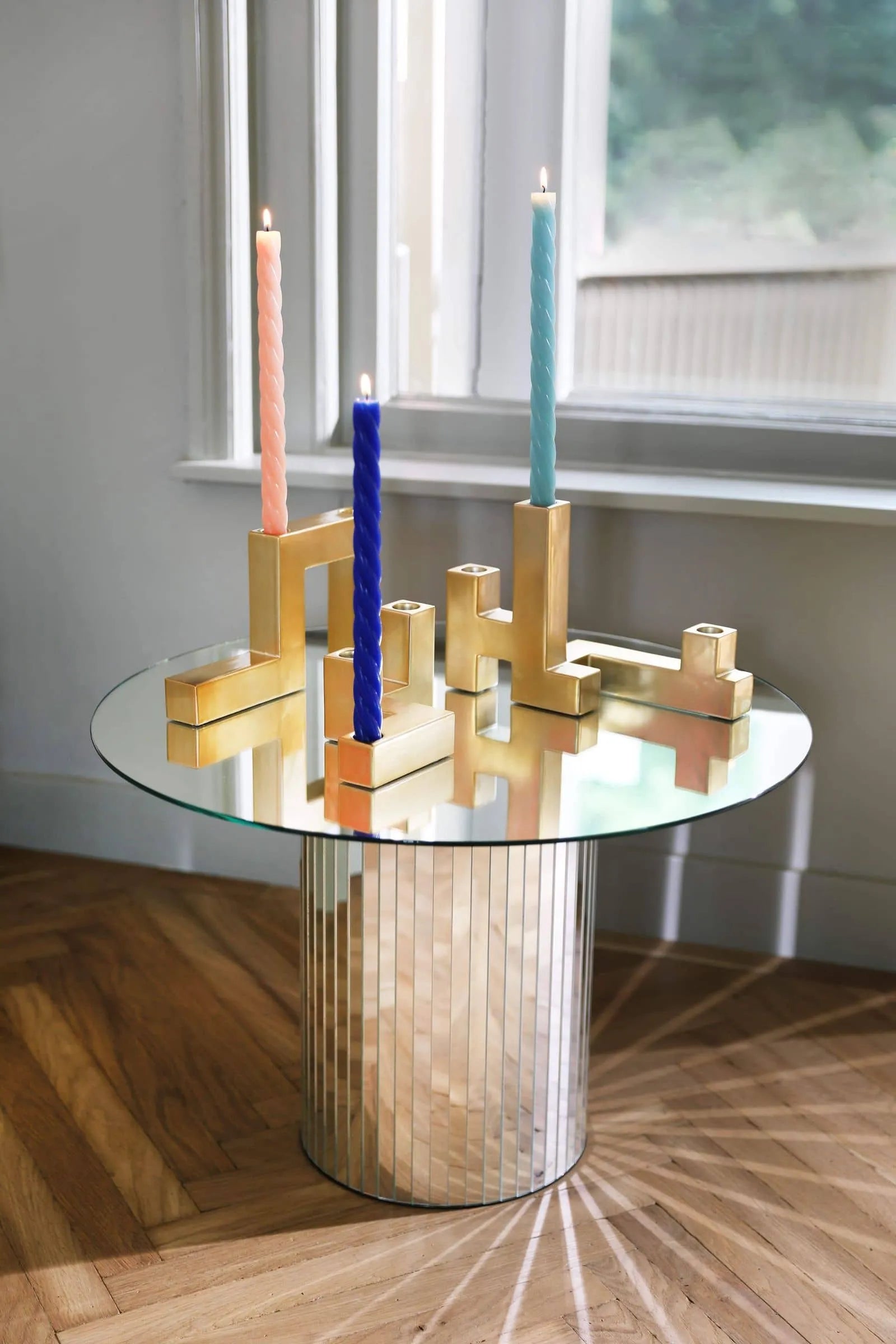 A round glass table displays gold holders with &klevering Marshmallow Candles (set of 5) in pastel pink, blue, mint green, and peach. Sunlight from a nearby window casts soft shadows on the parquet floor.