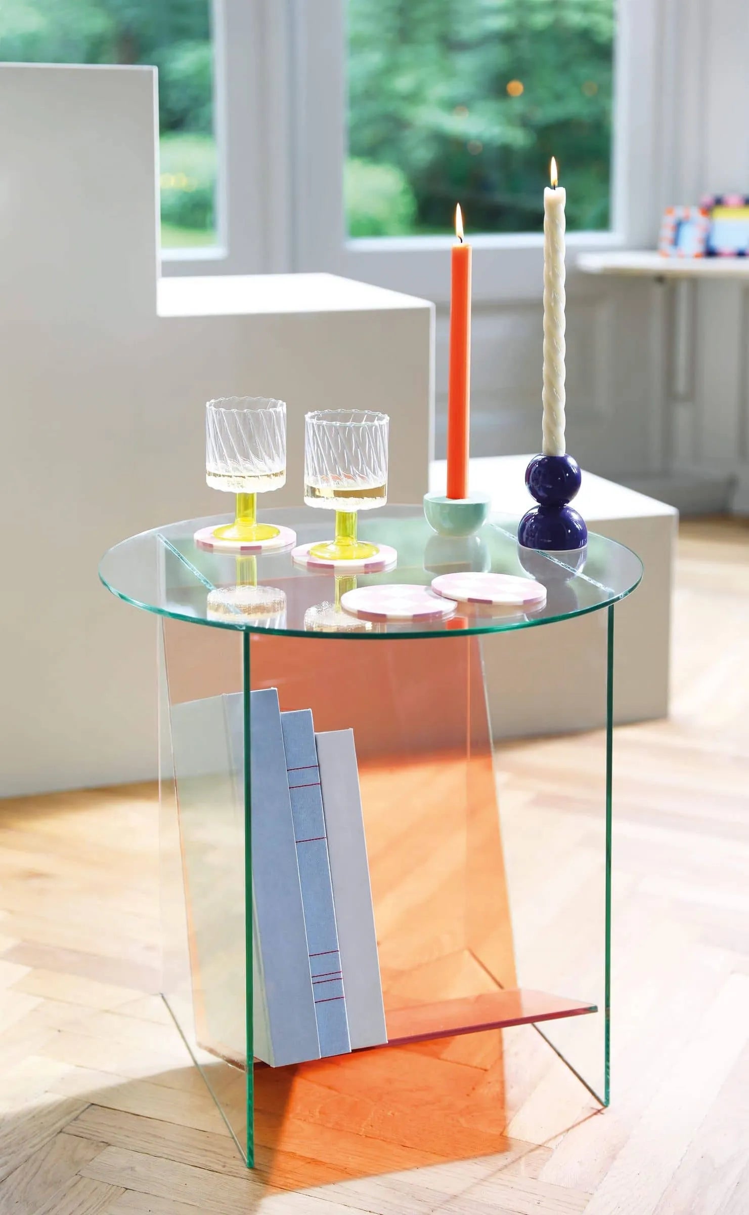 A modern glass side table with orange accents displays two glasses with yellow bases, a few books below, and the &klevering Marshmallow Candle, Set of 5 by &klevering. The background features large windows and minimalistic white furniture.