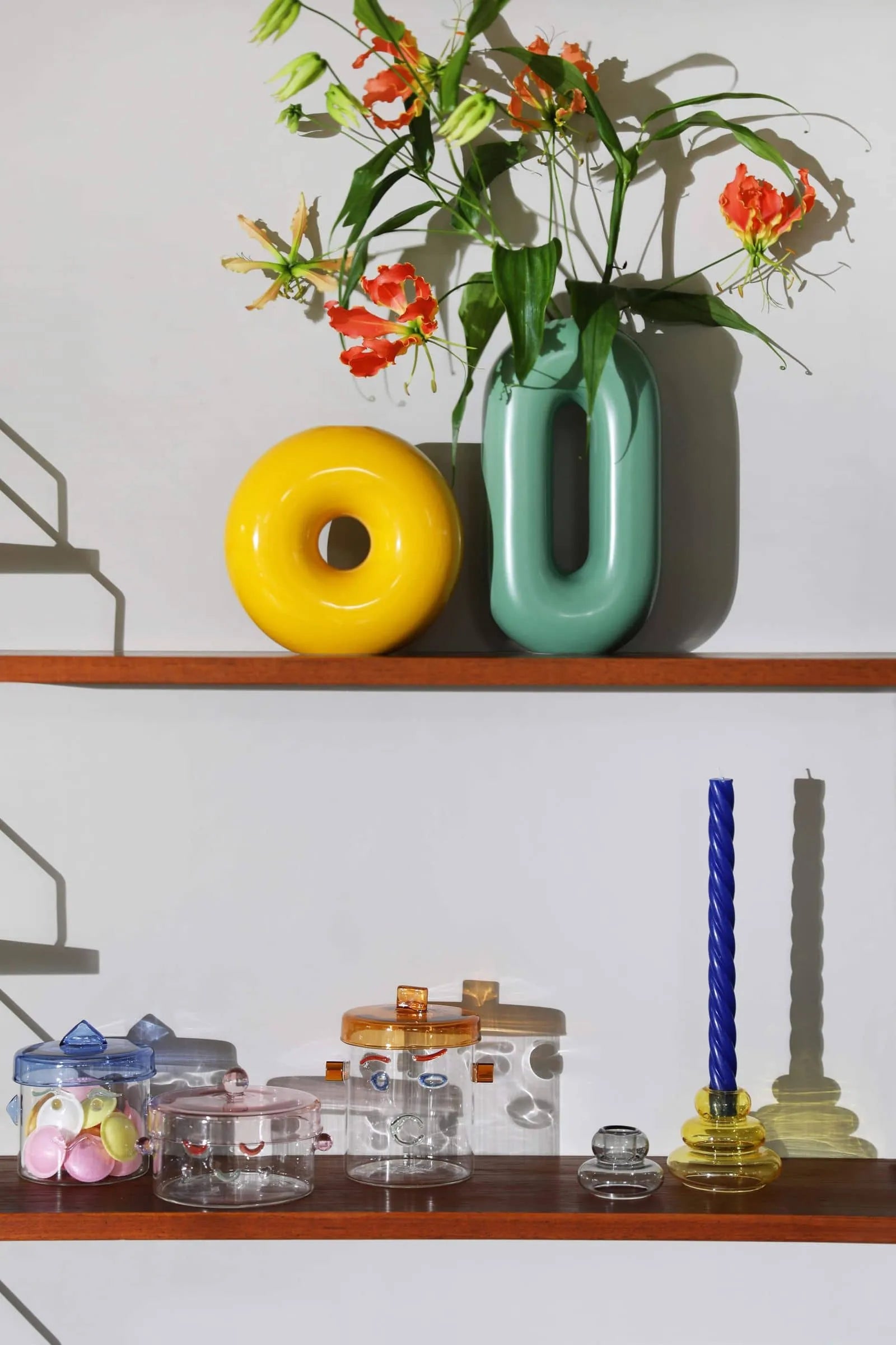 A shelf displays orange flowers in a green vase, a yellow donut sculpture, glass containers, colorful objects, and &klevering Marshmallow Candle set of 5 casting shadows on a white wall.