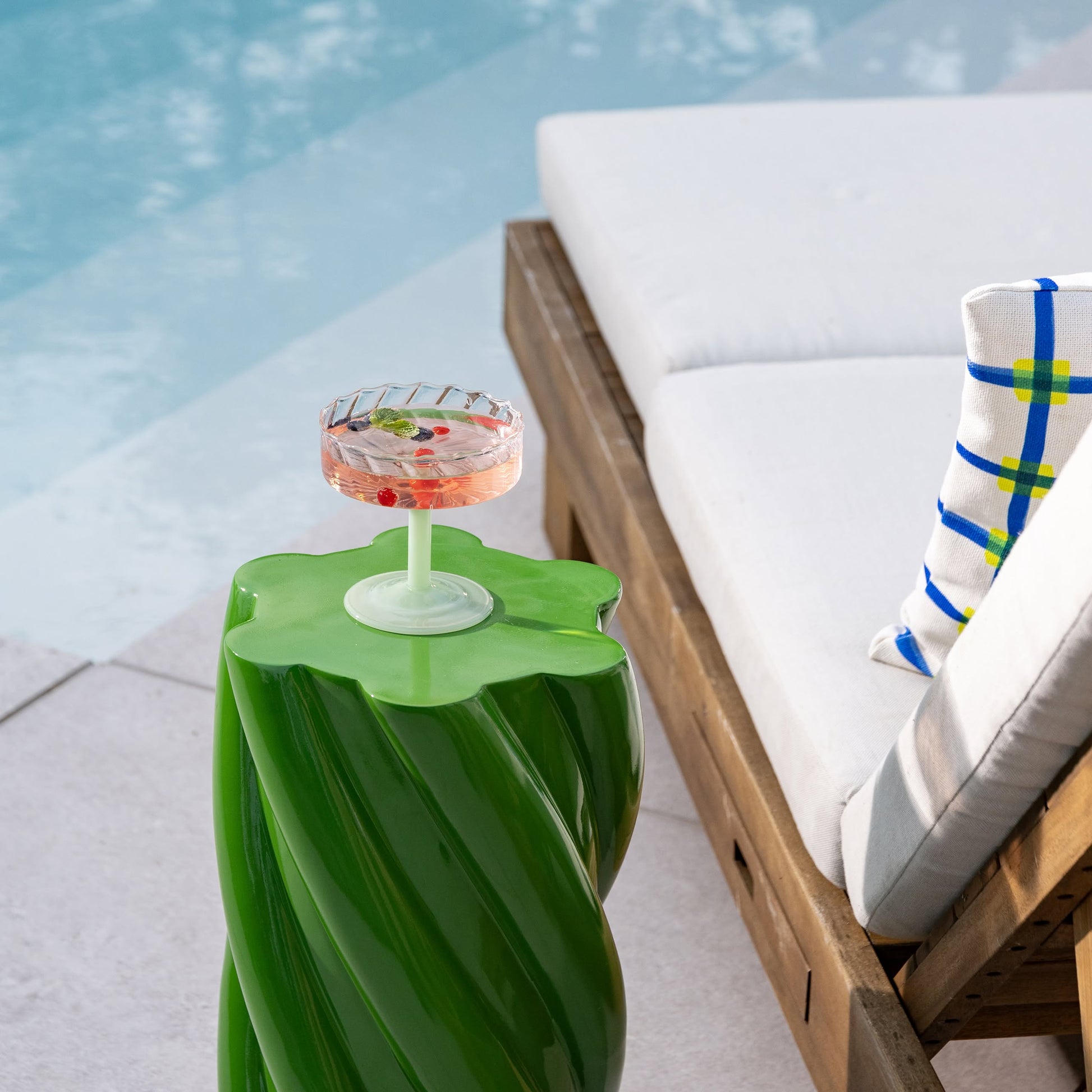 A cocktail with berries and herbs rests on the whimsical &klevering Marshmallow Pillar side table beside a wooden lounge chair with white cushions and a colorful plaid pillow, next to a pool.