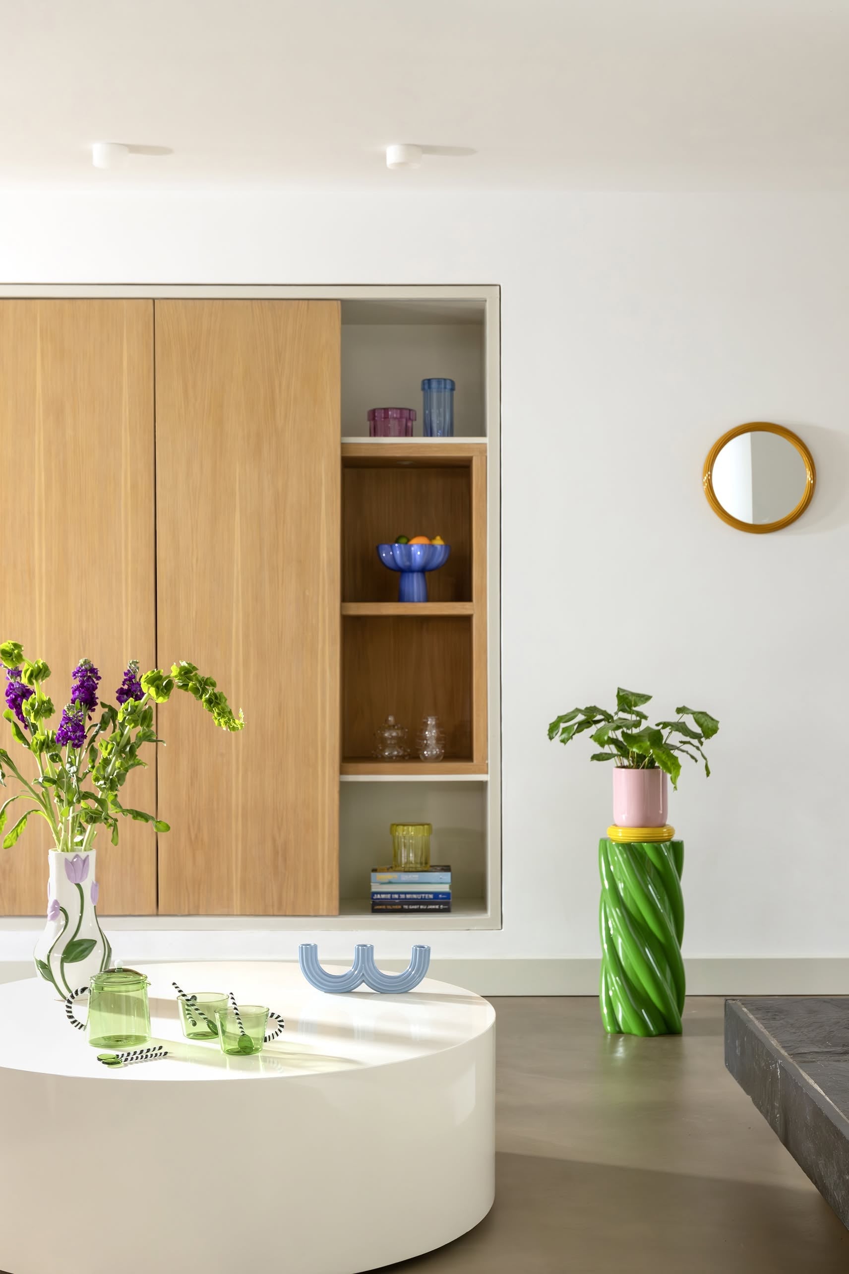 A modern living space featuring the &klevering Marshmallow Pillar, a white oval coffee table, playful design accents, colorful flower vases, a green spiral plant stand, built-in shelves with glassware, and a round mirror on a white wall.