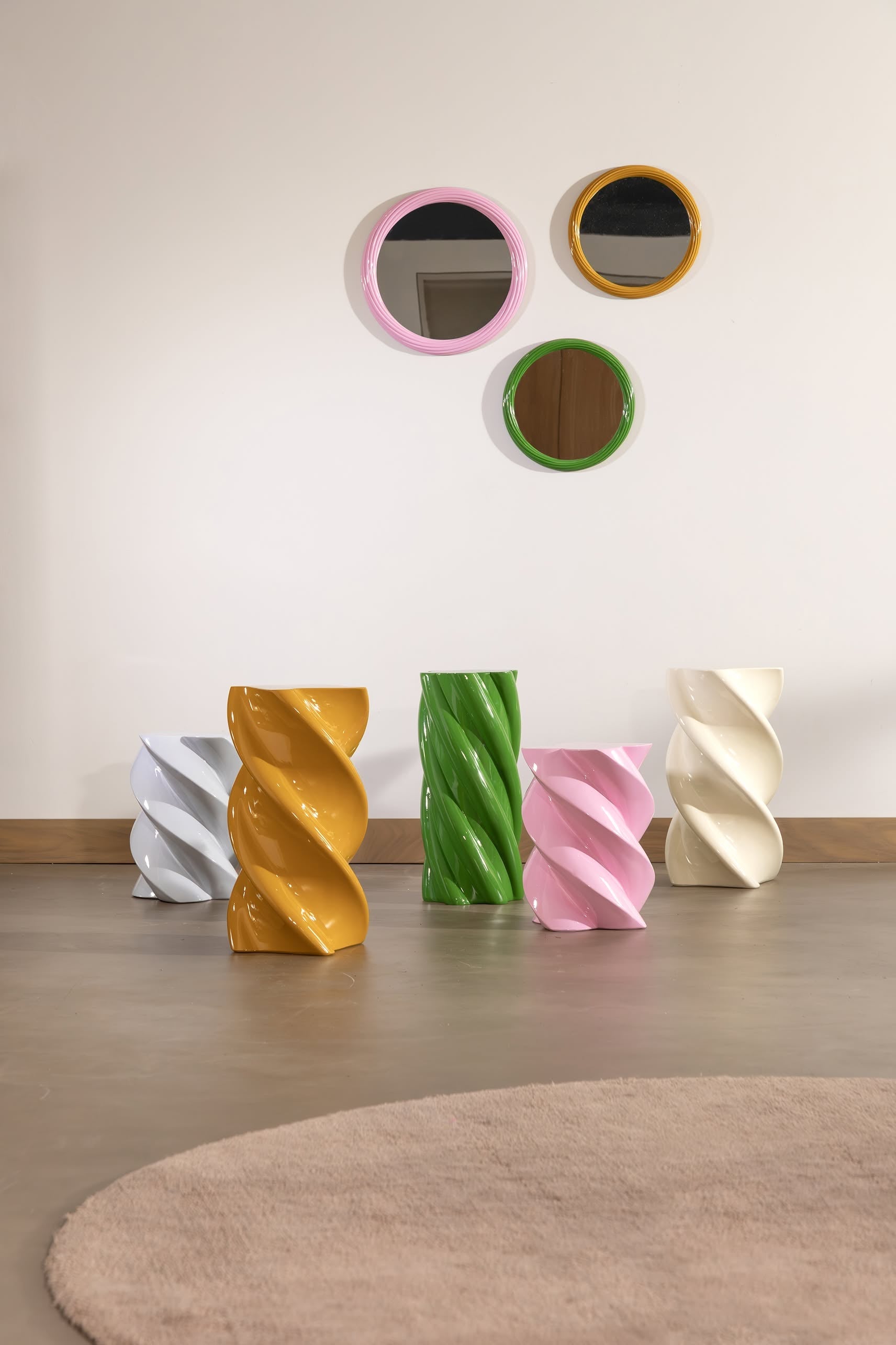 A minimalist room features five &klevering Marshmallow Pillar stools in white, gold, green, pink, and cream under three round mirrors with pink, gold, and green frames; a beige rug accents the floor.