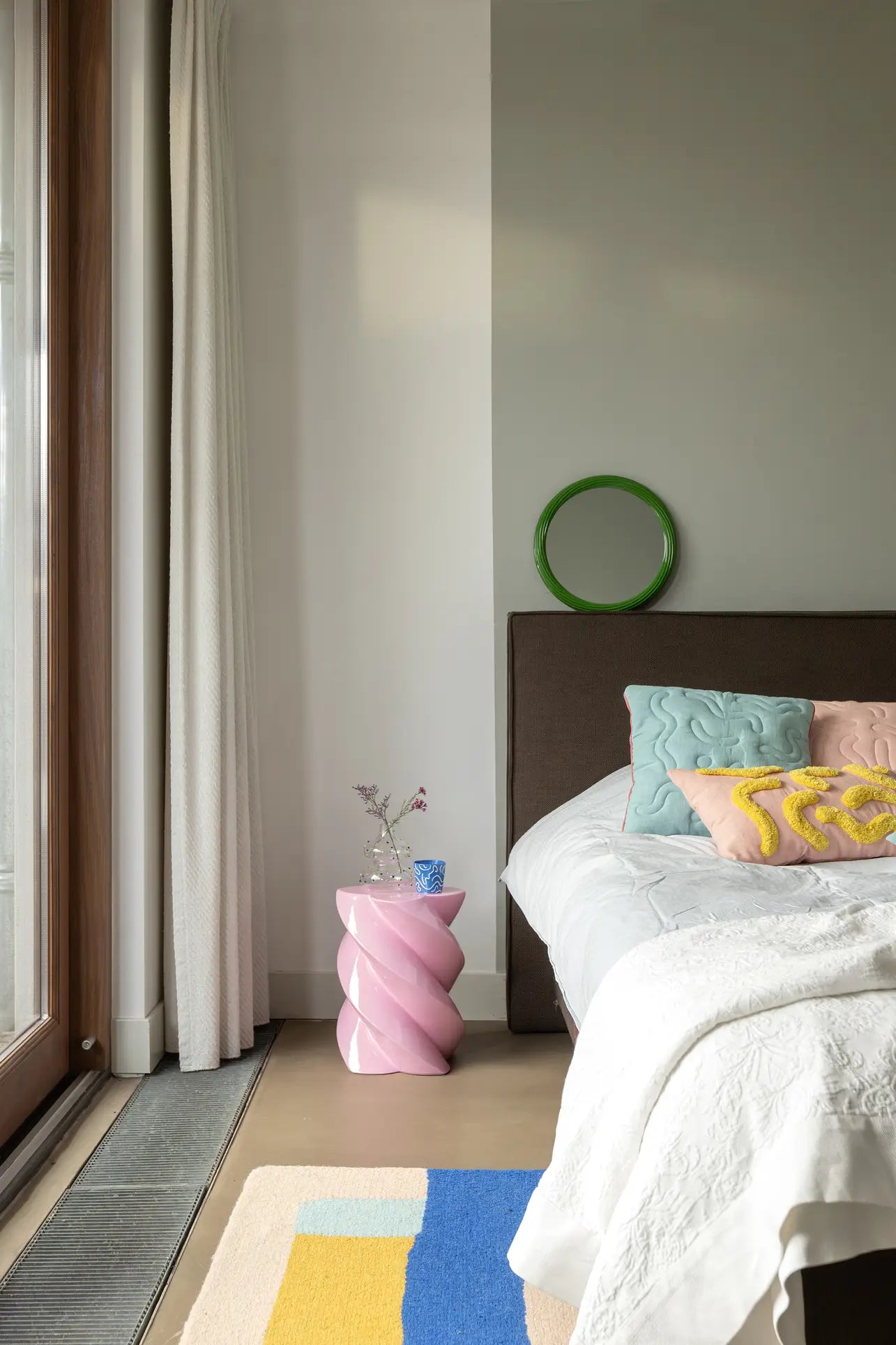 A modern bedroom with a bed adorned with pastel pillows and a white quilt, a whimsical &klevering Marshmallow Pillar decorative table topped with a vase and glass, and green circular wall decor above the gray-brown headboard by the window.