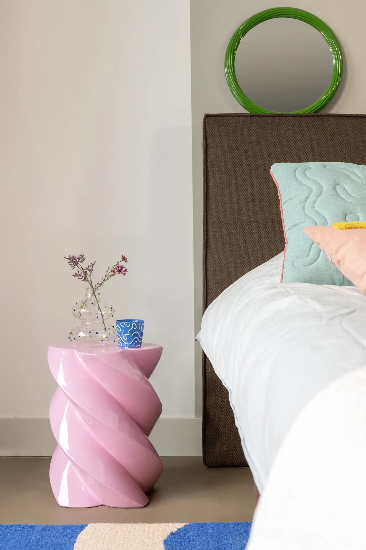 A modern bedroom features a white bed with blue and yellow pillows, brown headboard, and a decorative &klevering Marshmallow Pillar used as a side table for flowers and a blue cup. A whimsical round green-framed mirror adds the finishing touch.