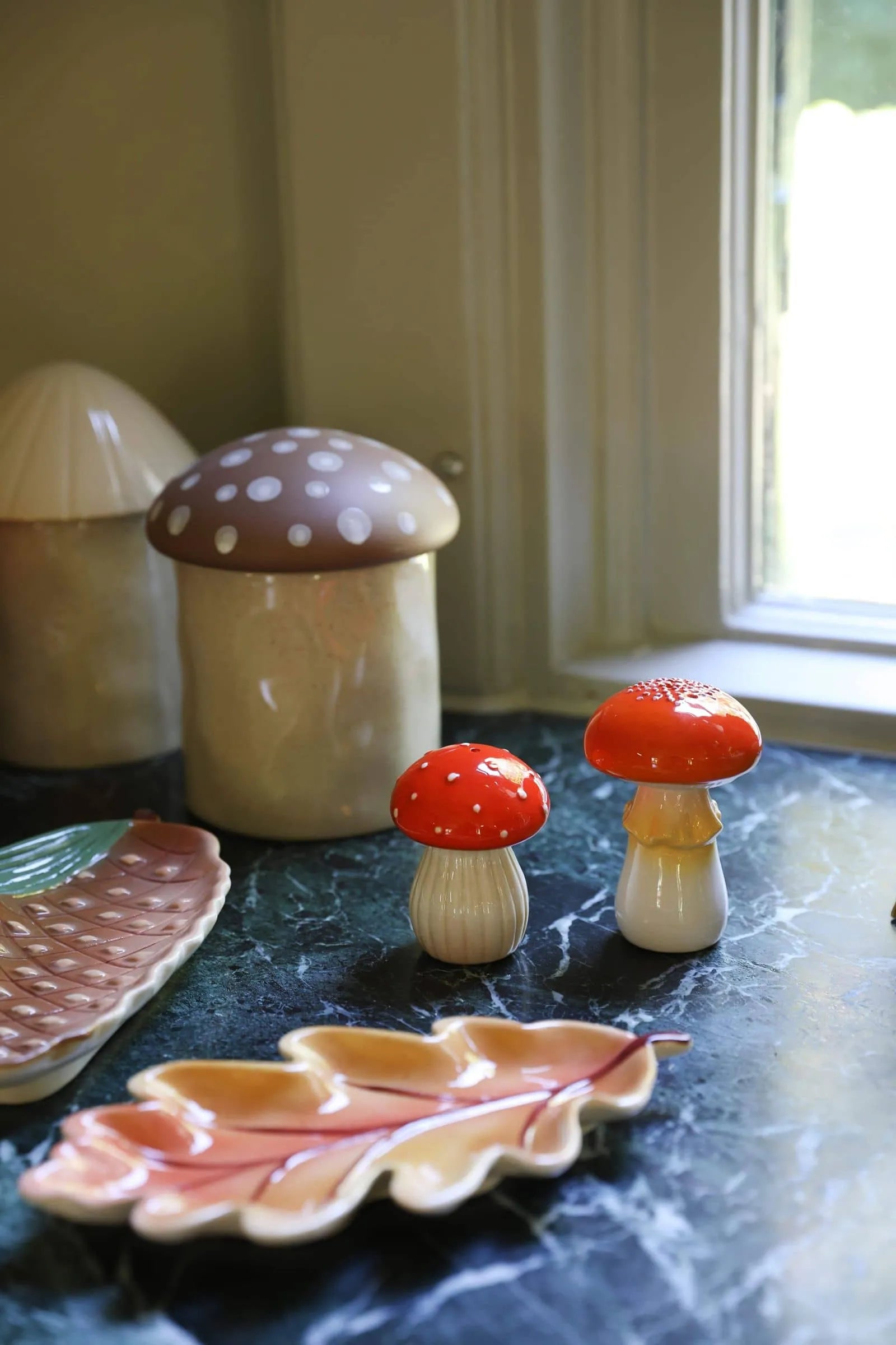 The &klevering Mushroom Salt & Pepper Set, featuring red and beige caps, sits on a green marble countertop by the window. Leaf-shaped ceramic dishes from &klevering add charm to this cozy kitchenโs dining experience.