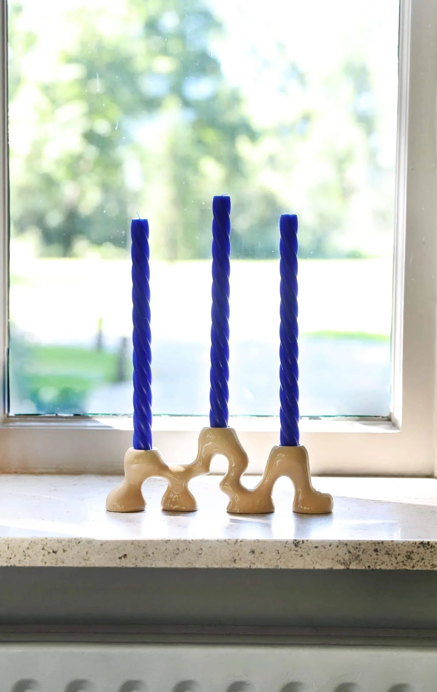 Three blue twisted candles are displayed in the &klevering Onda Small Candle Holder, by &klevering, featuring organic curves, set on a windowsill with a softly blurred outdoor view in the background.
