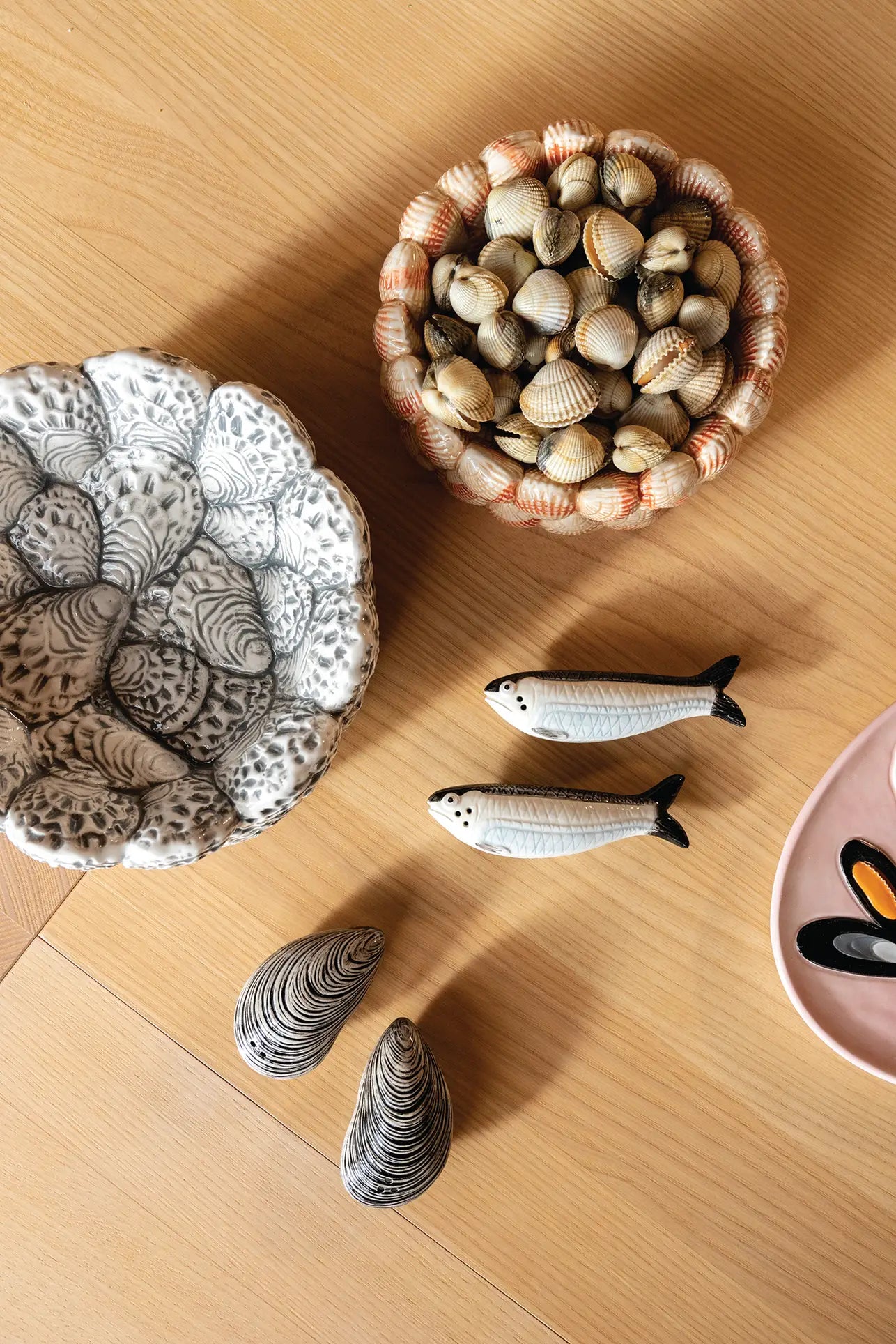 Ceramic shells, a bowl of seashells, two fish figurines, and the &klevering Oyster Bowl by &klevering with mussel designs are arranged on light woodโideal for serving appetizers or delighting oyster lovers.