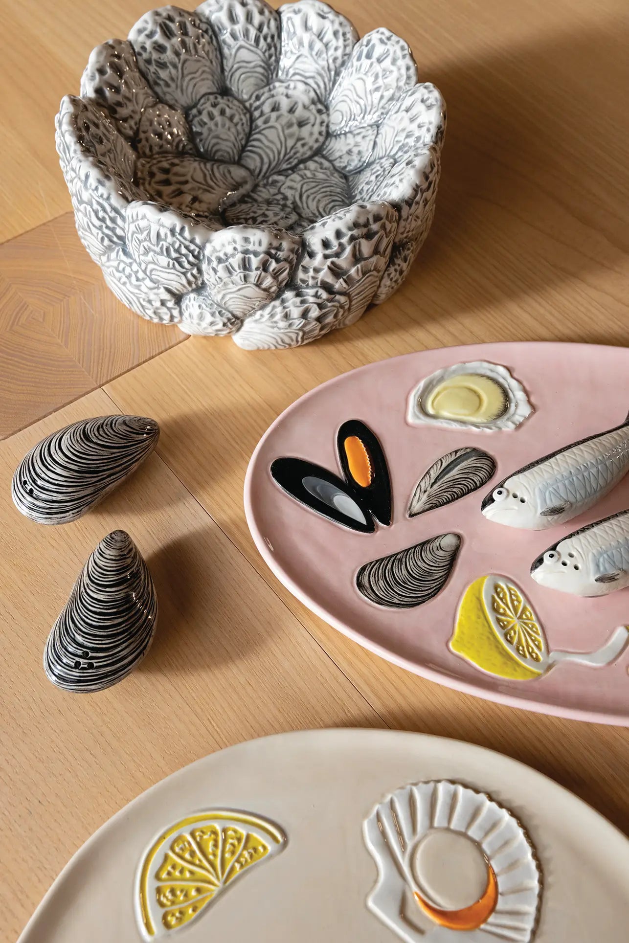 Ceramic dishware on a wooden table, including the detailed &klevering Oyster Bowlโperfect for appetizersโa pink plate with fish, mussels, and lemon designs, two mussel-shaped items, and a cream plate with lemon and shell motifs for oyster enthusiasts.
