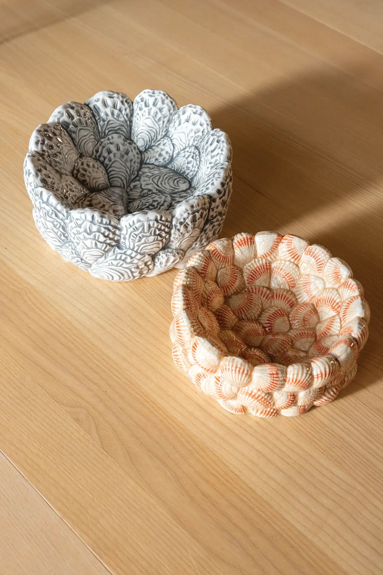 Two &klevering Oyster Bowls rest on a light wooden surfaceโone white with gray patterns, the other off-white with orange shell motifsโideal for appetizers or oyster lovers.