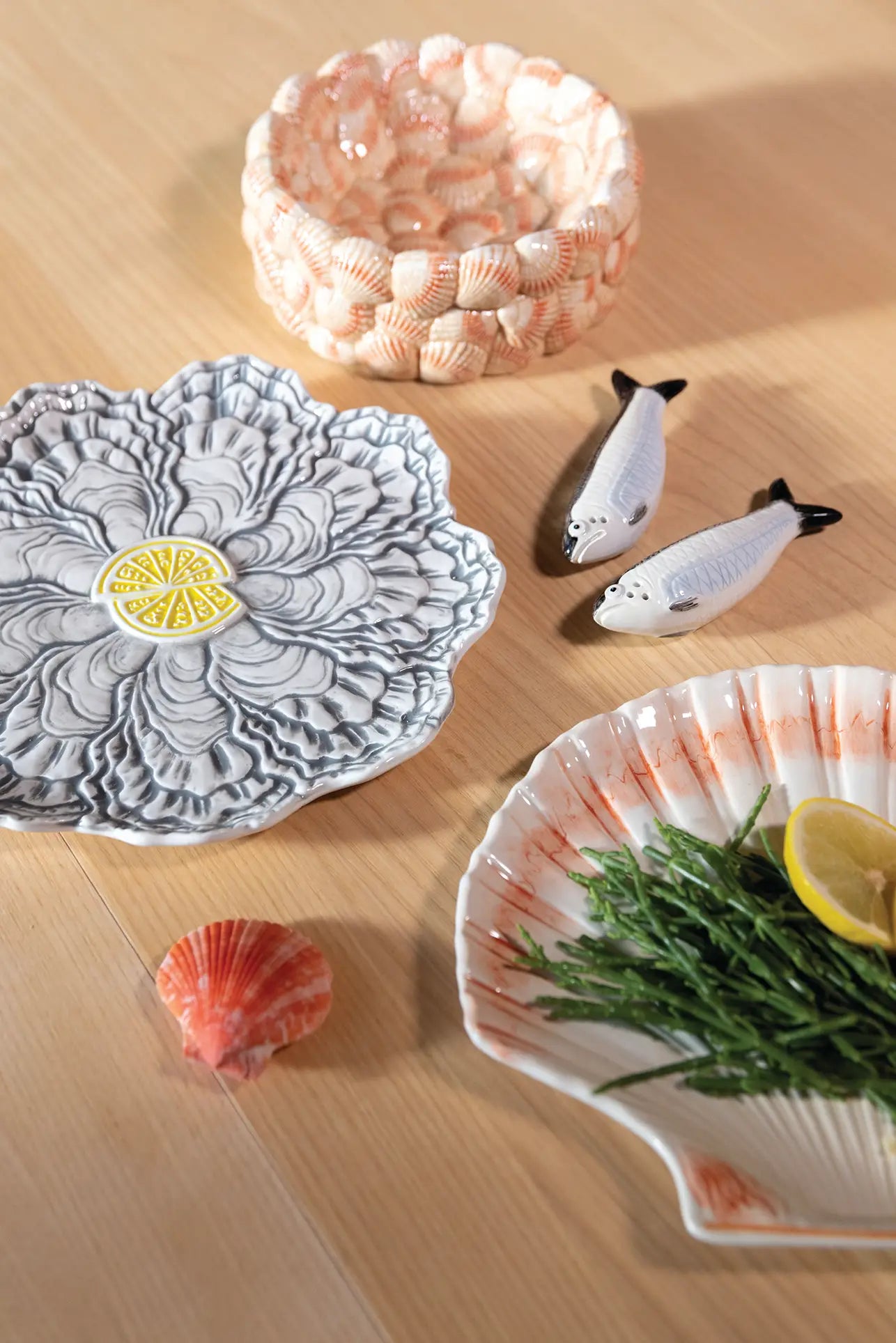 Ceramic seashell and flower-shaped plates, one styled for oysters with lemon and greens, a pink seashell bowl, two fish salt and pepper shakers, and an &klevering Oyster Plate by &klevering on a light wood surface.