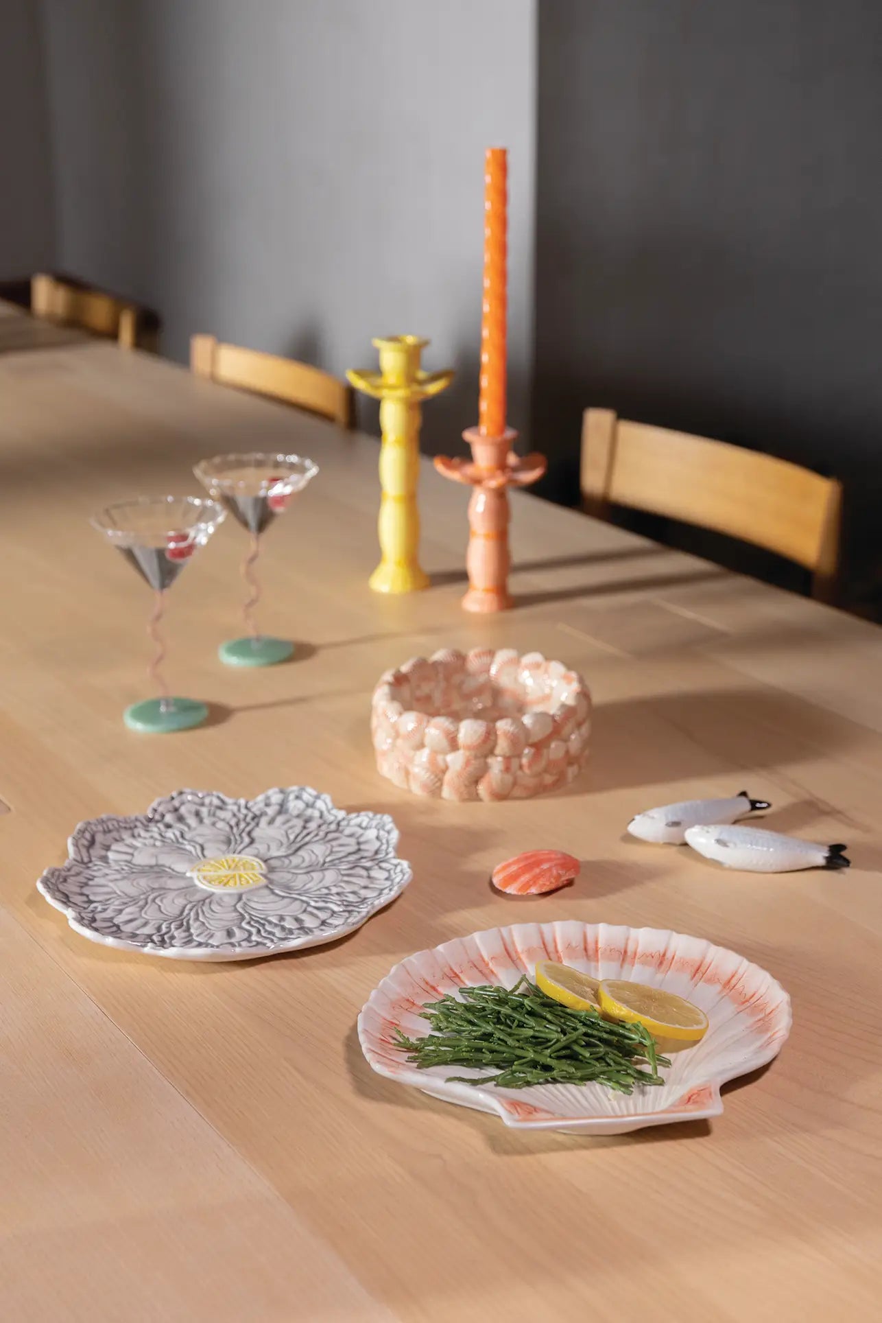 A wooden dining table set features two &klevering Oyster Plates—ideal for oyster lovers—arranged with greens, lemon slices, bird-shaped ceramics, a seashell, a round basket, wavy wine glasses, and tall yellow and orange candlesticks.