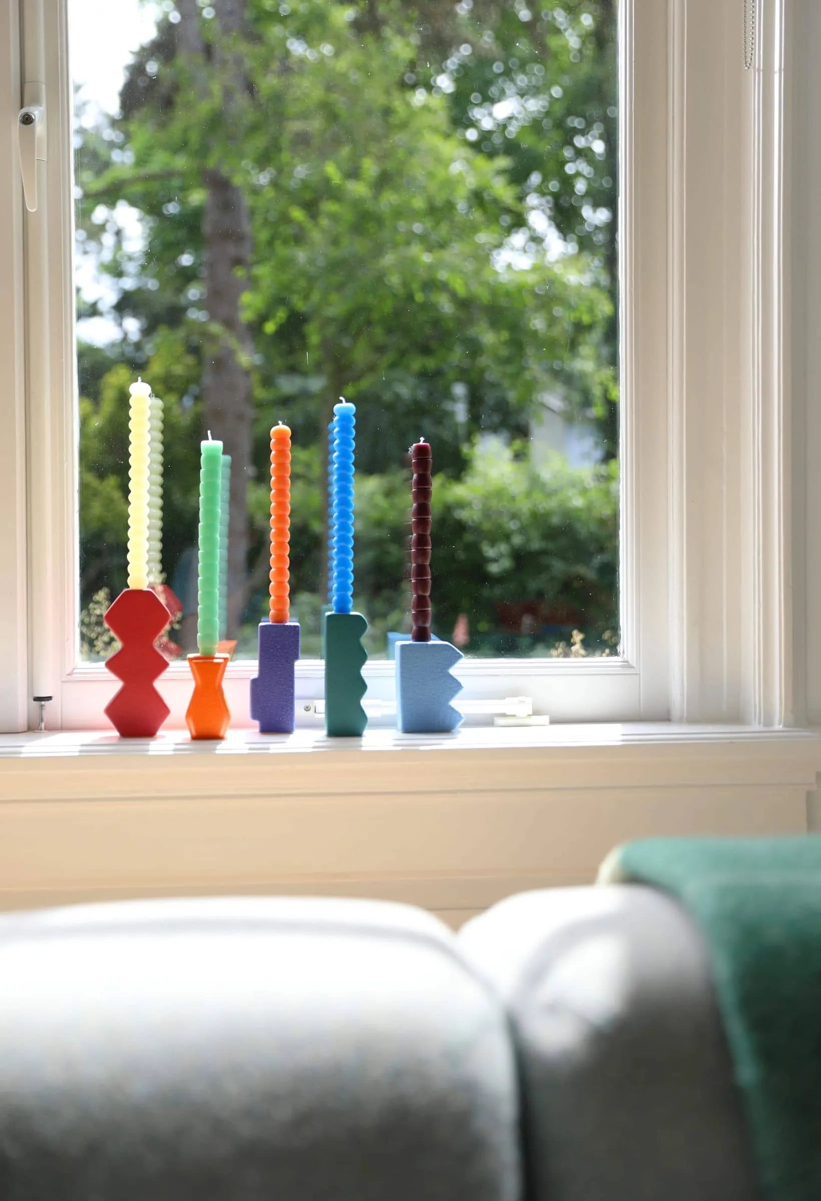 Set of 6 &klevering Pebble Candles in unique colors stand on colorful candle holders atop a white windowsill. Sunlight and green trees are visible through the window, with part of a couch in the foreground.