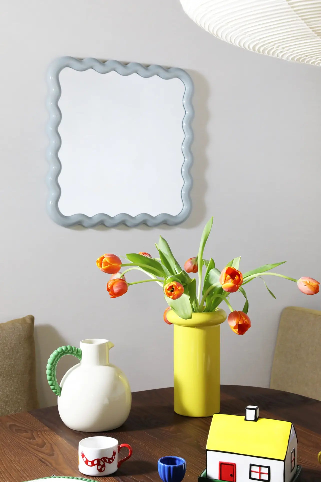 A modern dining table features a yellow vase of orange tulips, the &klevering Perle Jug, cups, and a small house ornament. A wavy-edged mirror above cushioned chairs adds elegance for perfect summer dining.