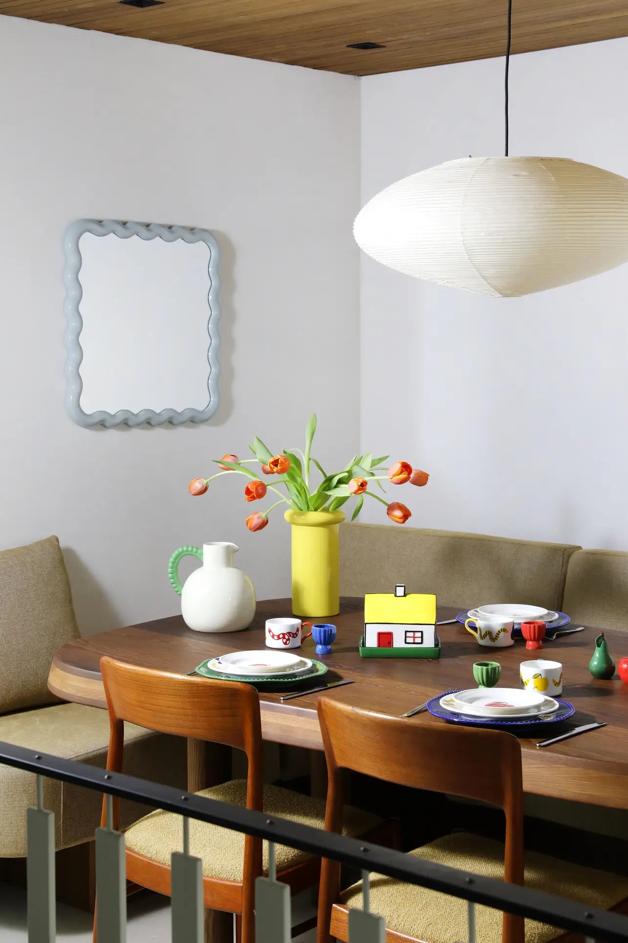 A modern dining area featuring a wooden table for four, beige bench and chairs, a &klevering Perle Jug with orange tulips, colorful décor, a wavy-edged mirror, and a large white pendant lamp—ideal for summer dining.