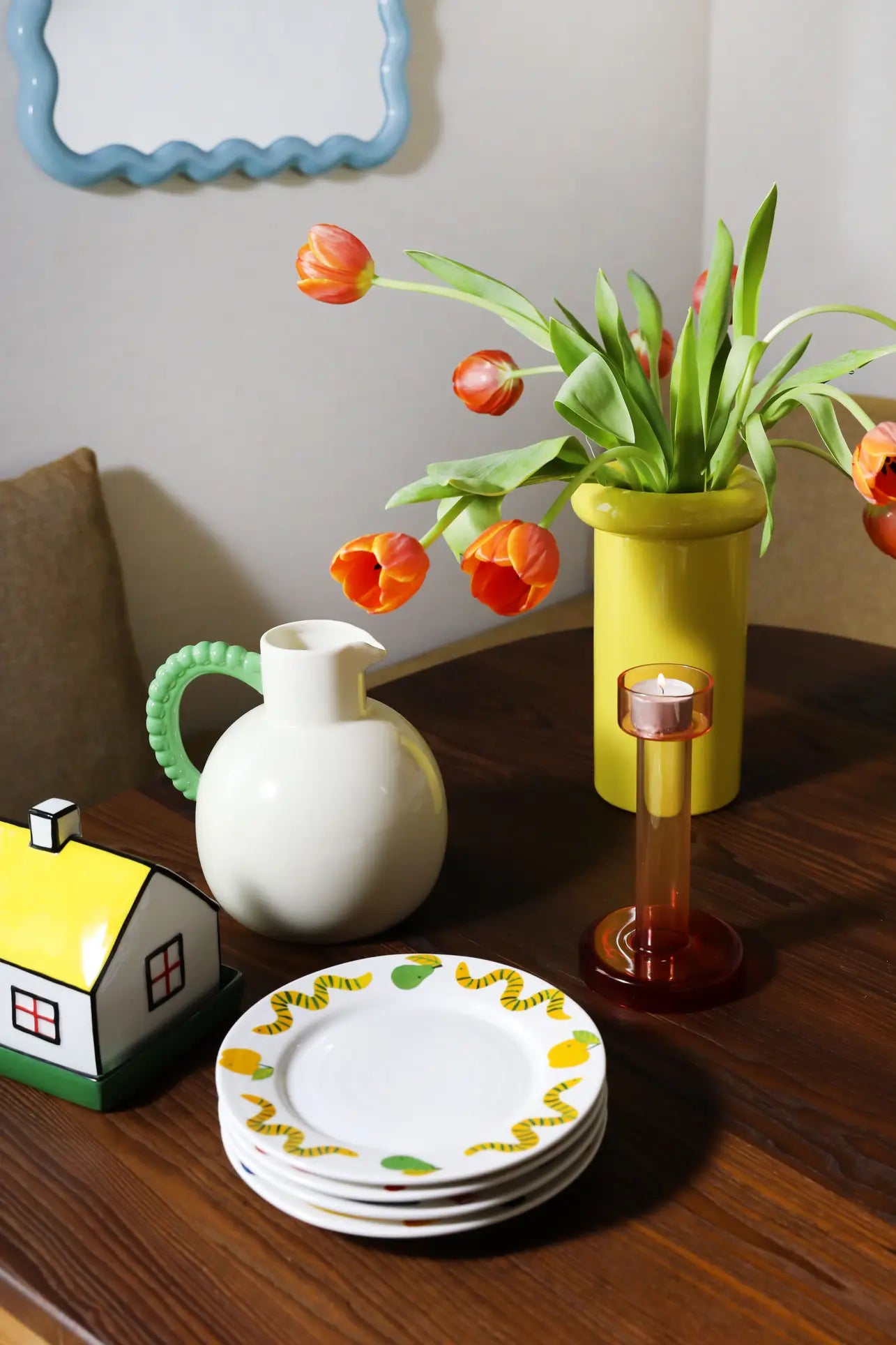 A wooden table with stacked plates, a house-shaped container, and the &klevering Perle Jug sits beside a tall yellow vase of orange tulips—an elegant summer dining setup. A wavy blue mirror in the background adds stylish charm.
