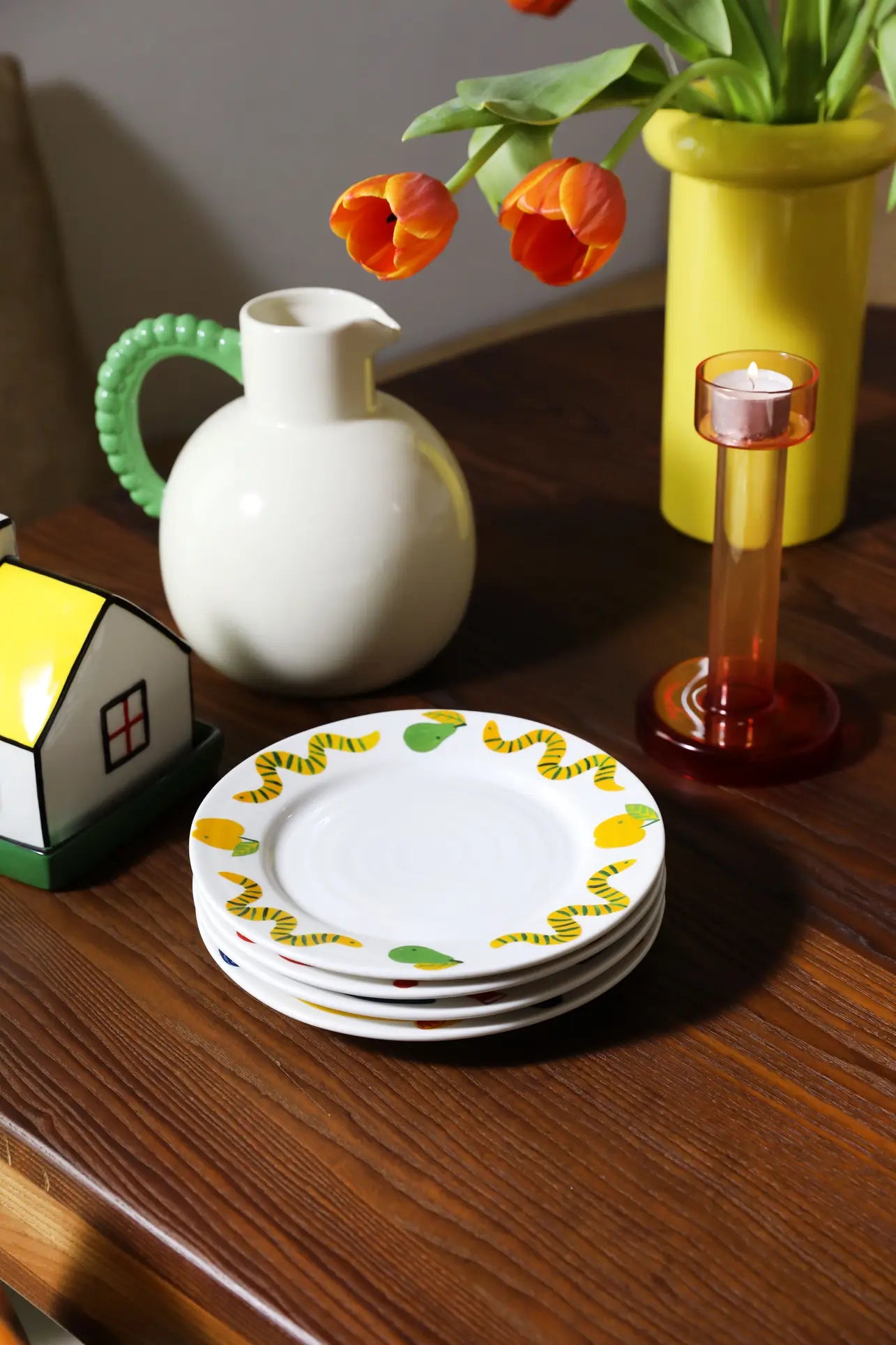 A stack of white plates with yellow and green designs sits on a wooden table next to the &klevering Perle Jug, an amber candle holder, a small house-shaped container, and a vase of orange tulips—perfect for elegant summer dining.