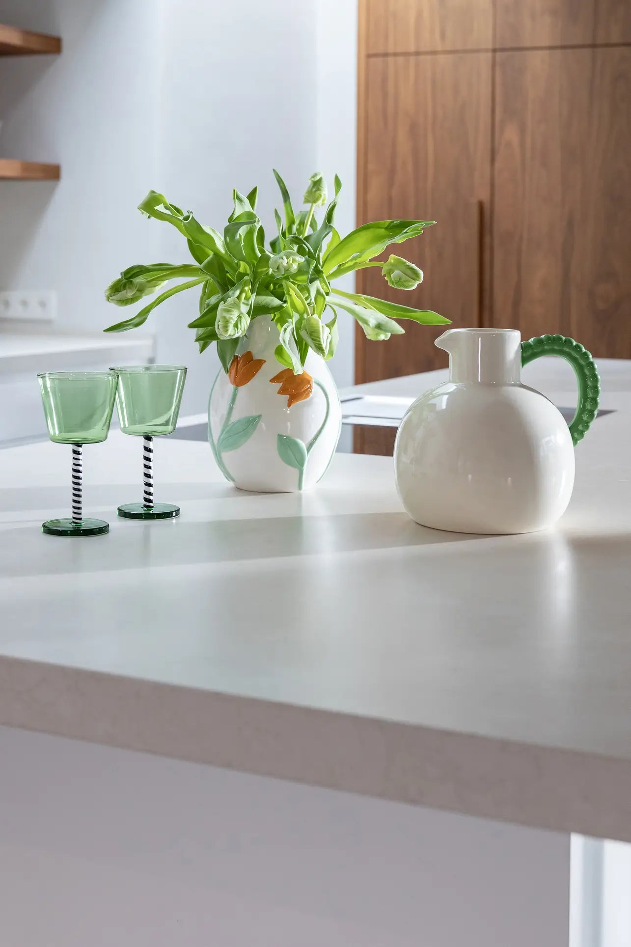 A modern kitchen countertop features two green glasses, a white vase with green leaves and orange flowers, and the elegant &klevering Perle Jug—perfect for summer dining—while wooden cabinets and shelves complete the background.