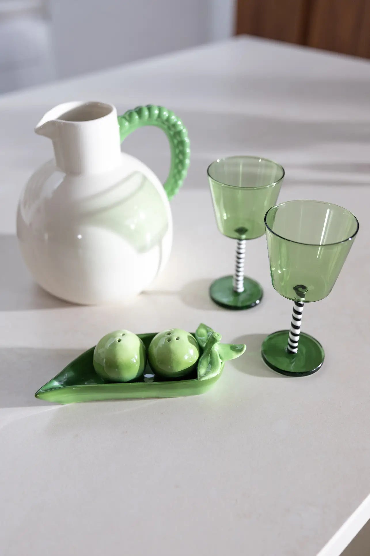 The &klevering Perle Jug with a green handle, two striped-stem wine glasses, and a pea pod-shaped salt and pepper dish create an elegant look perfect for summer dining on a light countertop.