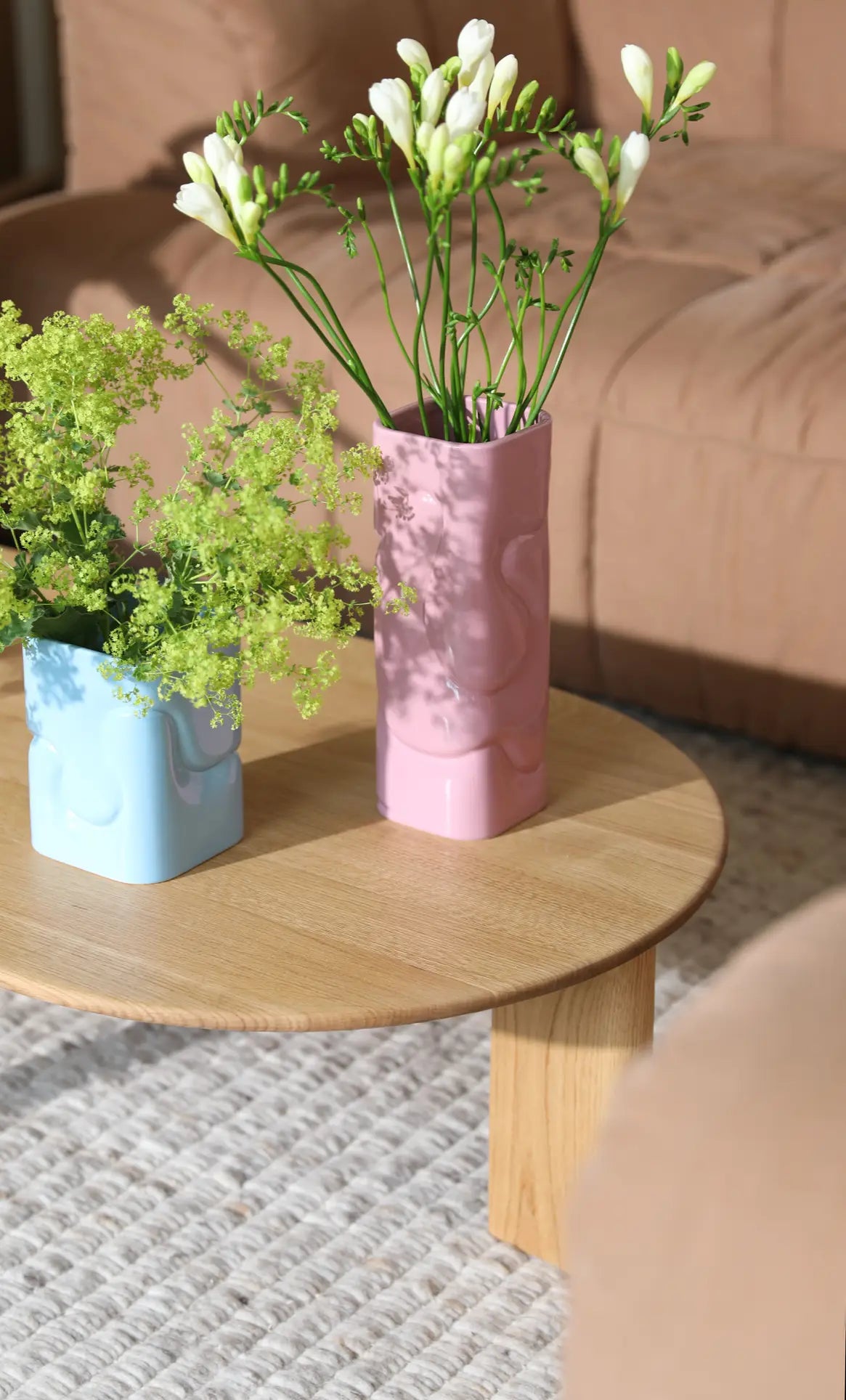 Two &klevering Puffy Vases, one light blue with green foliage and the other pink with white blooms, sit on a round wooden coffee table. A beige sofa and textured rug are in the background.