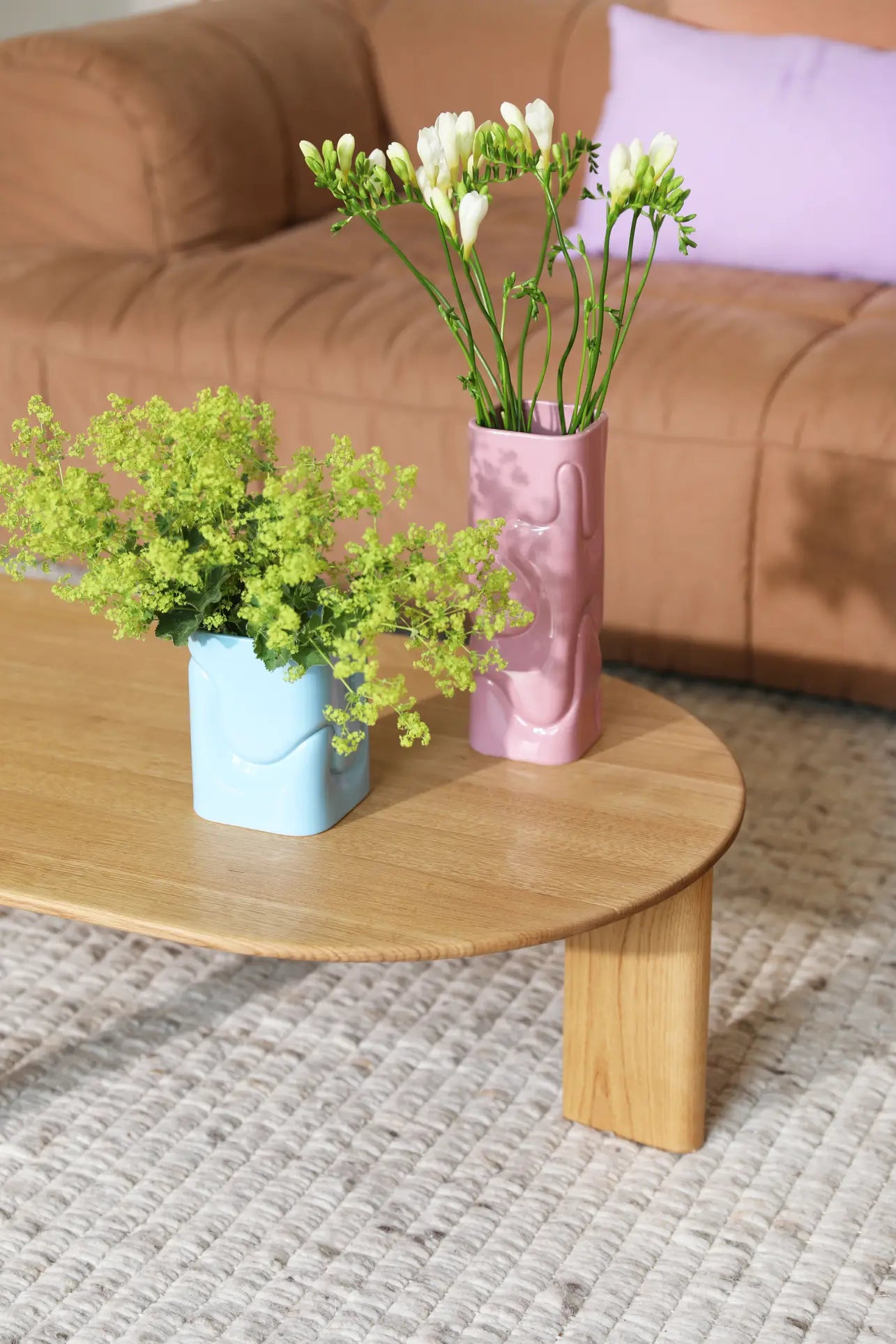 A wooden coffee table with an &klevering Puffy Vase and a pink vase with vibrant flowers sits on a textured rug in front of a tan sofa topped with a light purple cushion.