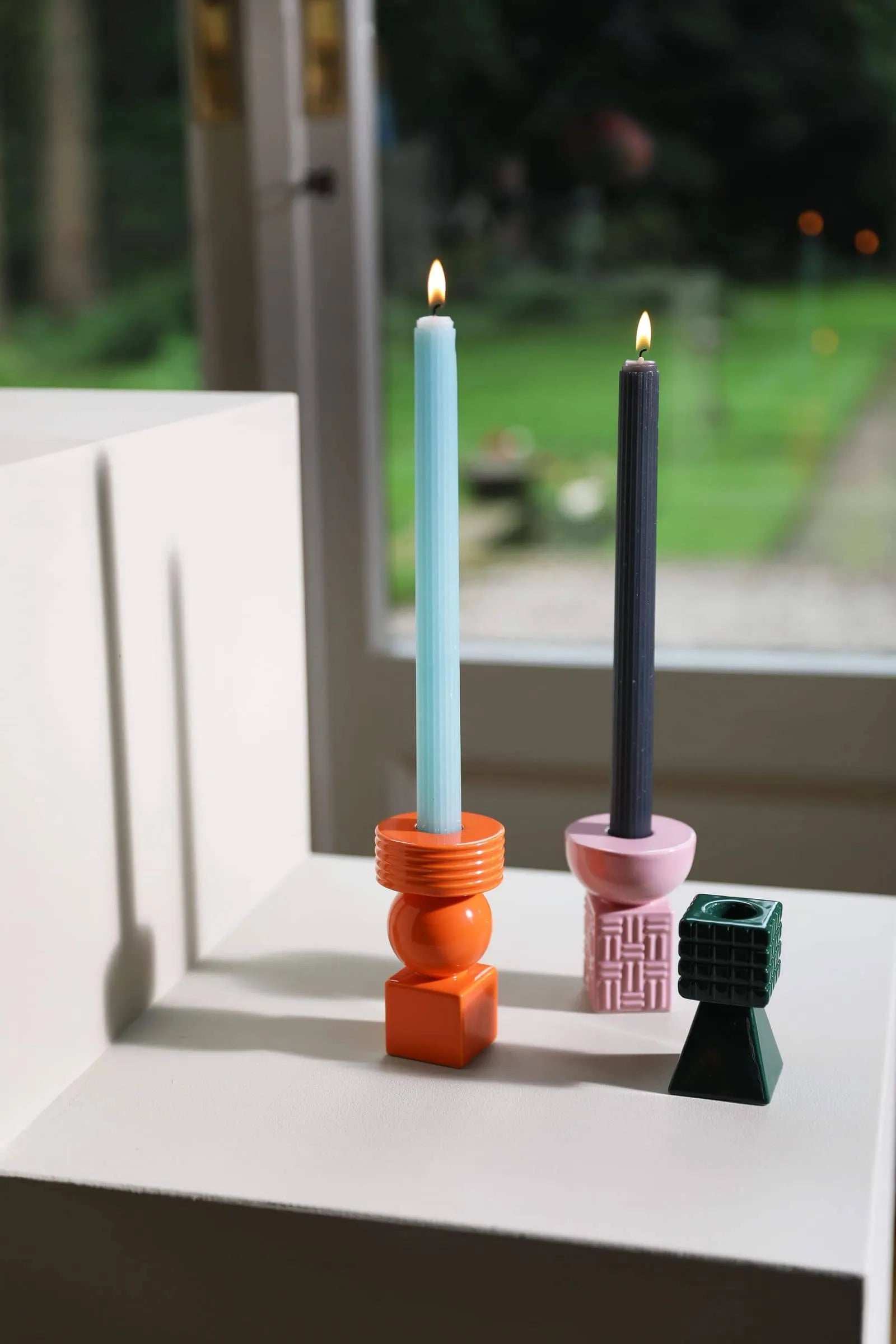 The &klevering Ripple Candle, Set of 5 by &klevering features handcrafted candles with vertical lined textures, shown here in vibrant orange and pink beside a small green geometric holder by a window overlooking a blurred garden.