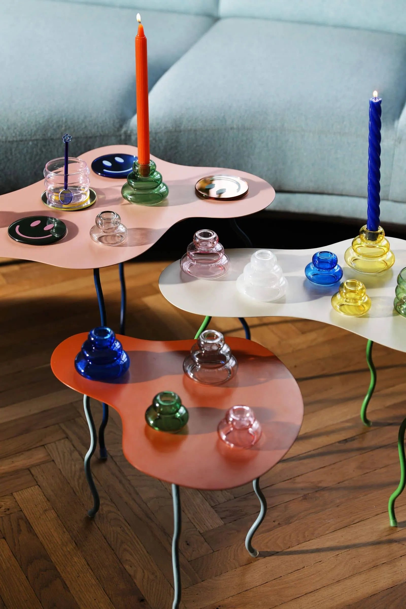 Three abstract, colorful side tables with wavy shapes display &klevering Ripple Candle, Set of 5—handcrafted candles with a unique vertical lined texture—on a wooden floor in front of a pale blue sofa.