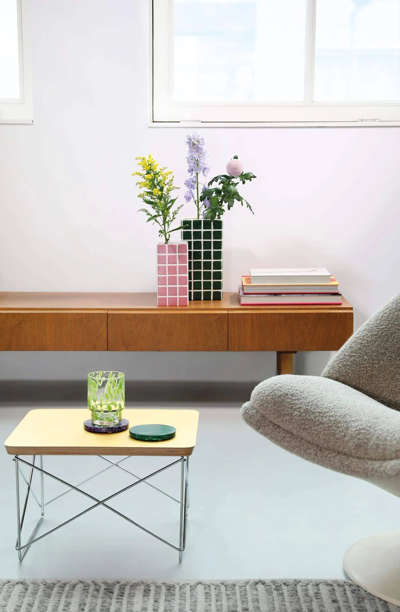 A modern living room features a mid-century wooden console displaying botanical treasures in the &klevering Tiled Vase Pink by &klevering, alongside stacked books. A light wood table with a green glass and coaster sits near a gray upholstered chair.