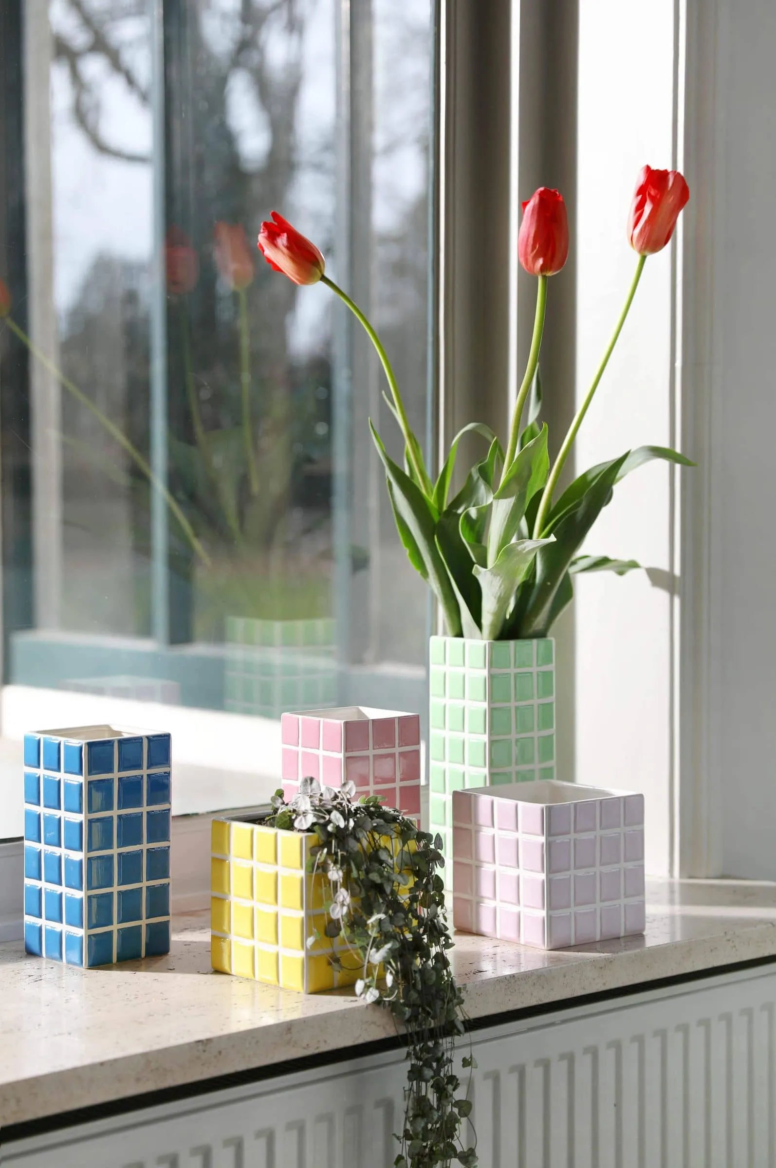 The &klevering Tiled Vase Pink from &klevering adds a candy-colored touch to the windowsill, its vibrant pink tile pattern catching the light as sunlight streams through the window.