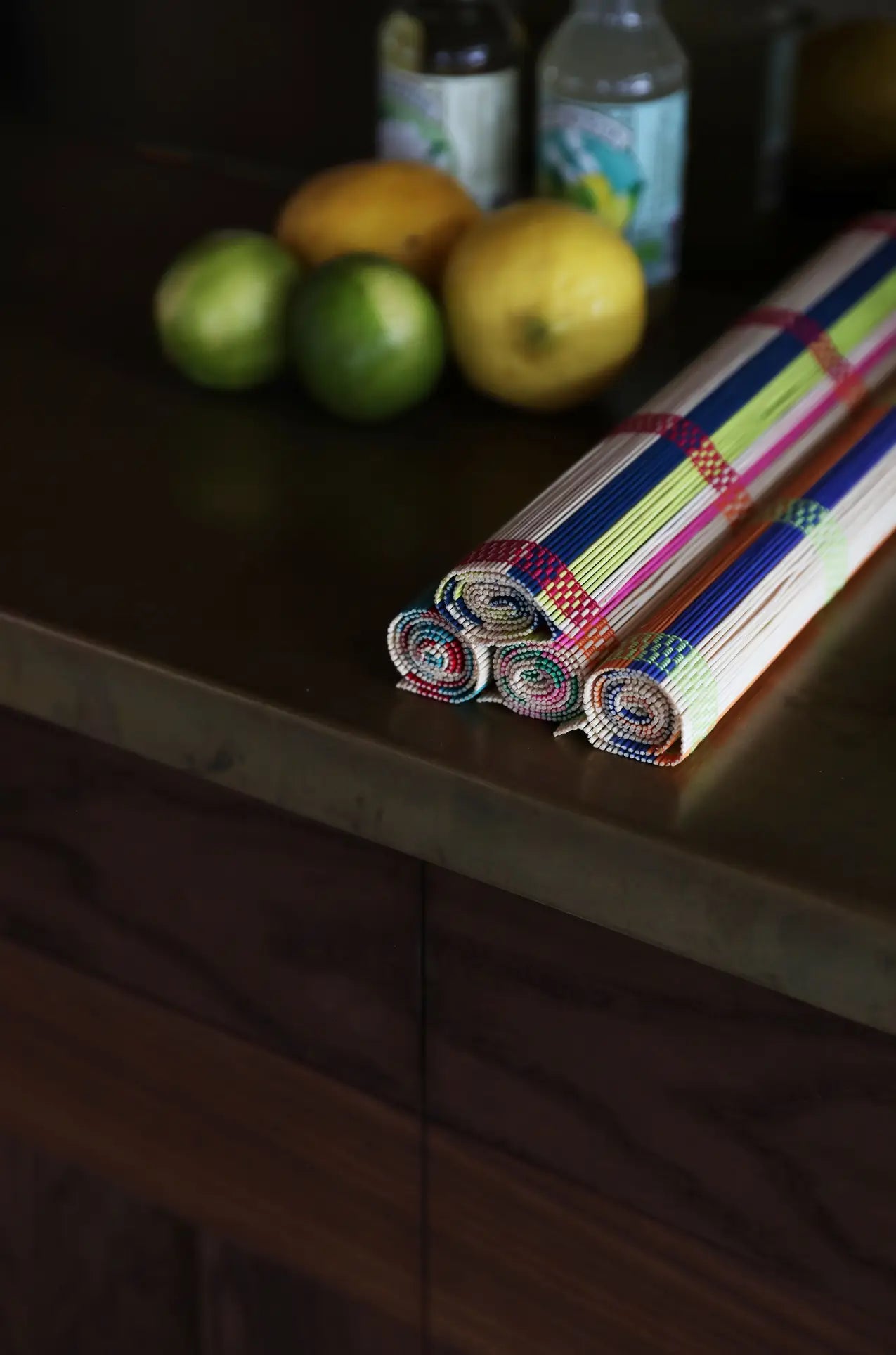 A set of three colorful &klevering Umami Placemats rests on a dark wooden countertop, with blurred lemons, limes, and bottles in the background.