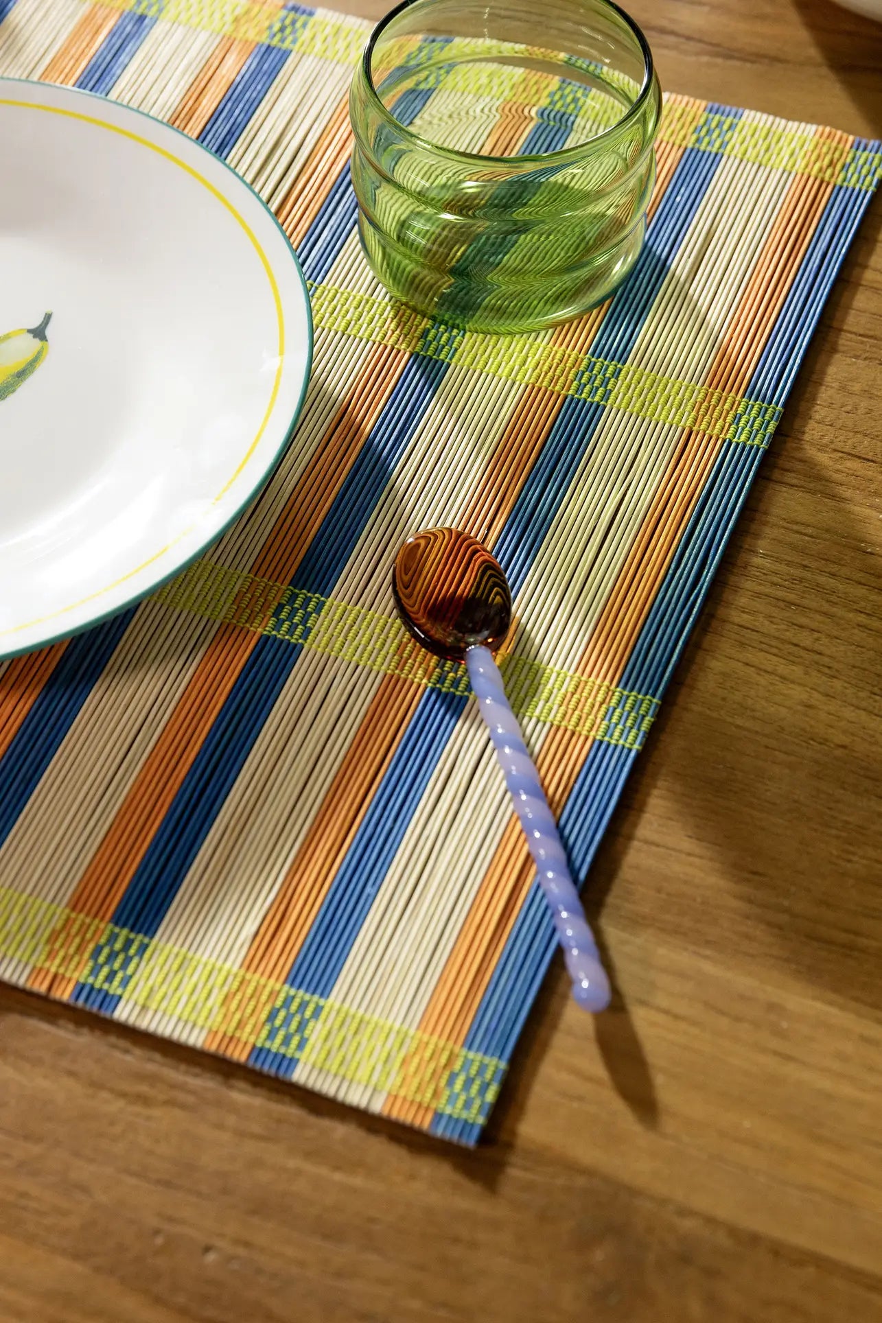 A twisted purple and white glass spoon rests on an &klevering Umami Placemat beside a green glass and a lemon-patterned plate, creating a vibrant dining setup with stylish pieces from &klevering.