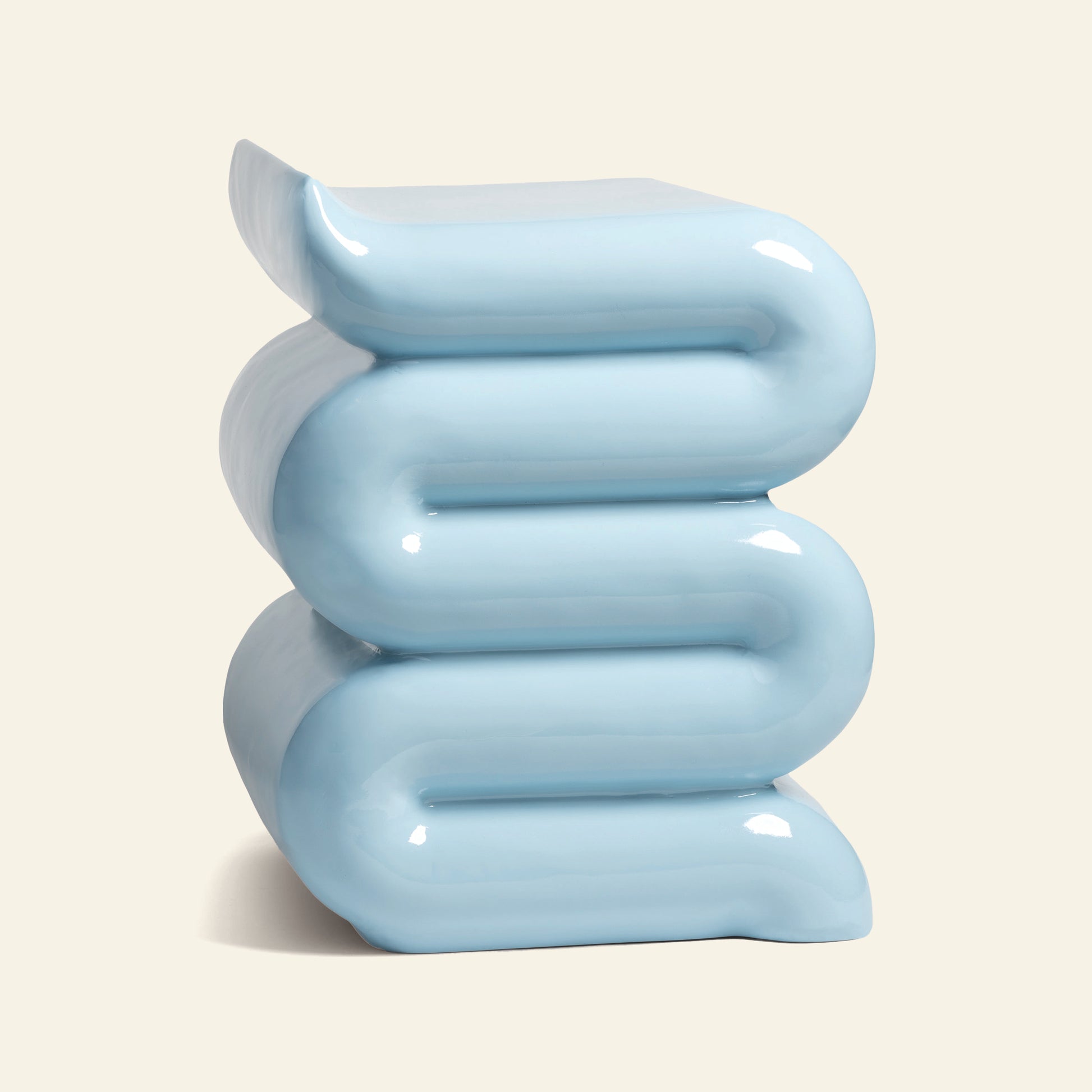 The &klevering Whip Pillar - Blue by &klevering is a glossy, pastel blue side table featuring a modern wavy, stacked design of three curved layers—an ideal accent for a cozy reading nook. Shown on a simple beige background.
