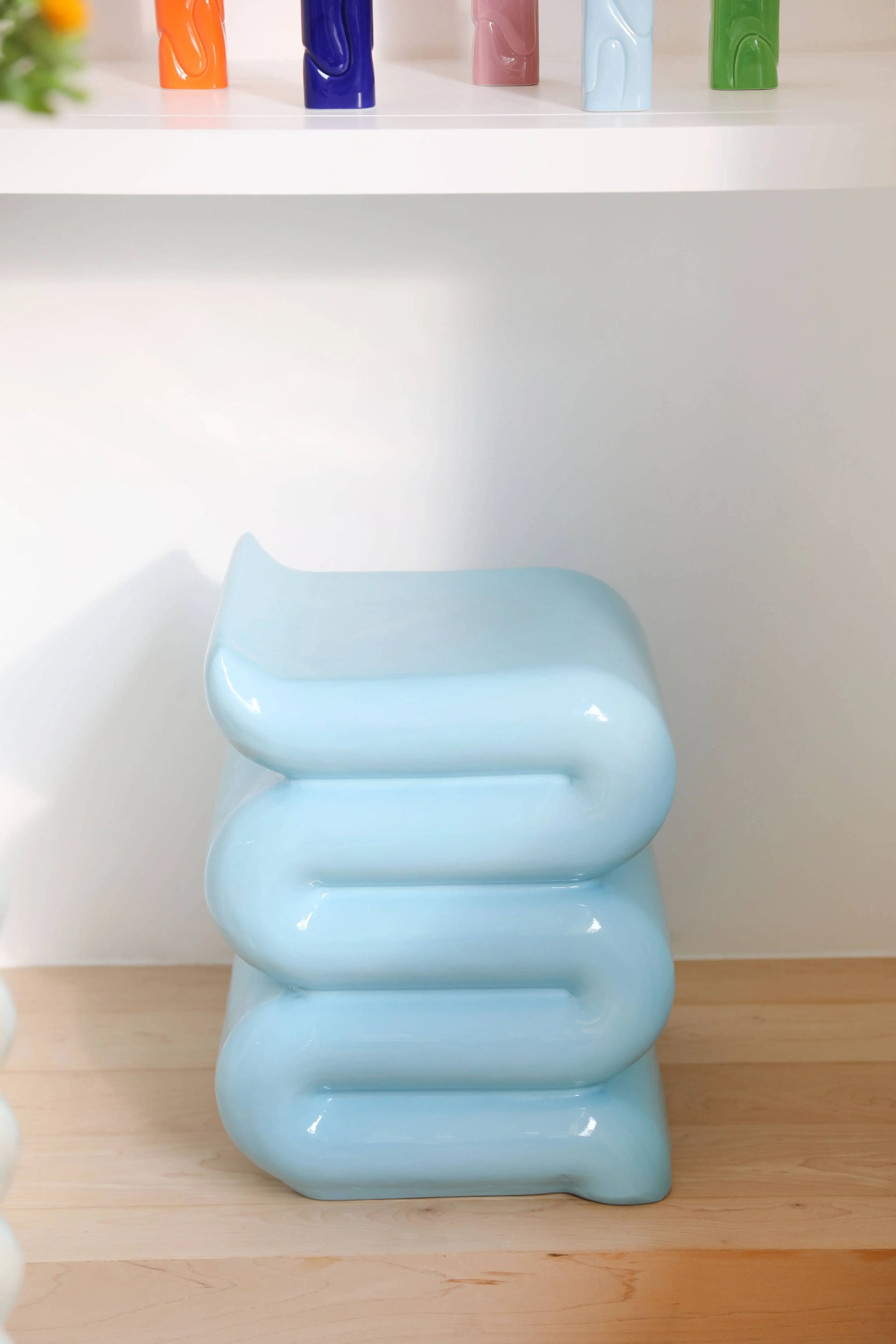 The &klevering Whip Pillar - Blue, a glossy, light blue modern stool with a curved, stacked design by &klevering, sits on a light wood floor against a white wall—ideal as a pastel side table for your reading nook.