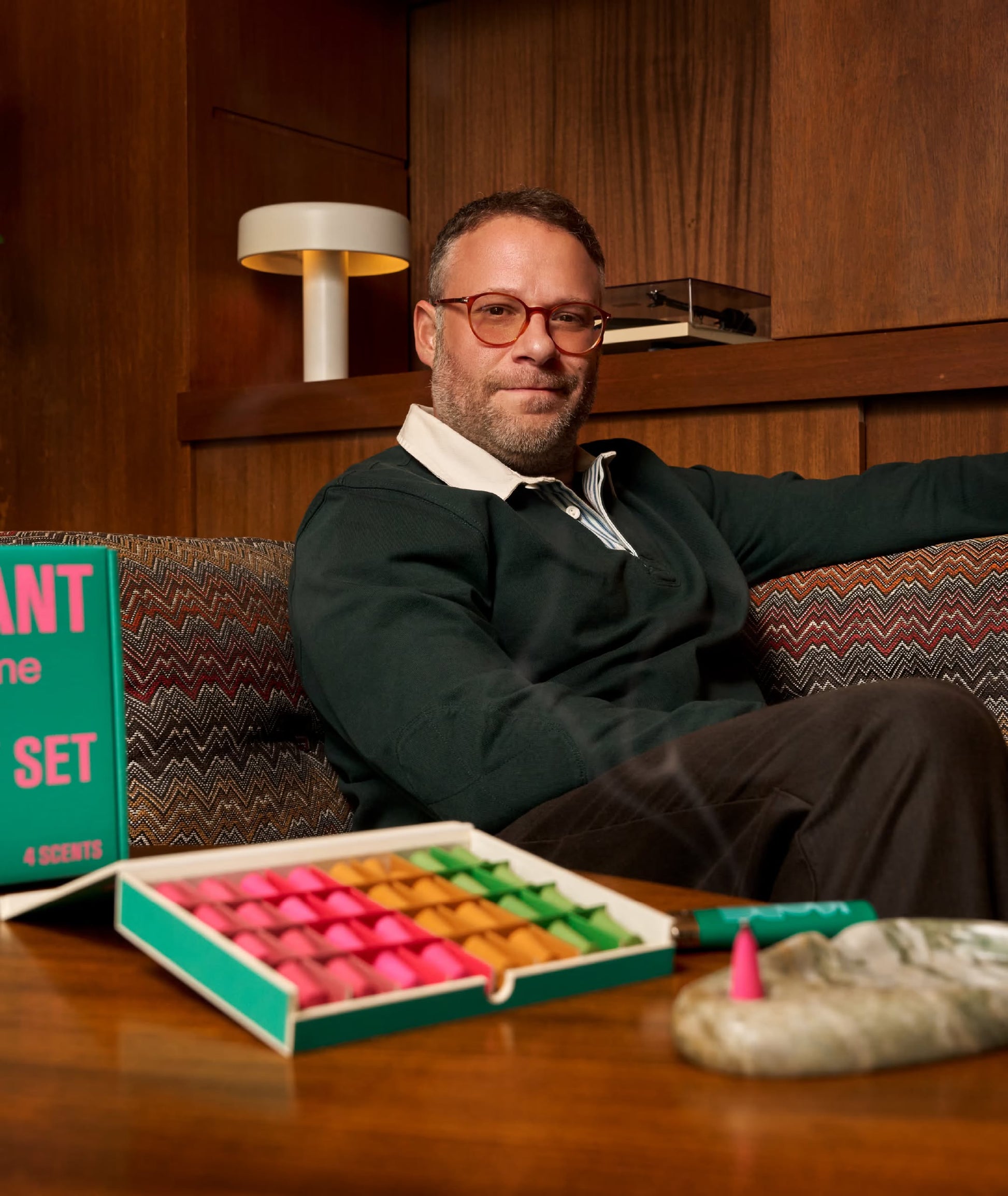 A man with glasses sits on a patterned sofa beside the ripple+ Houseplant Ash & Scent Set, with one cone burning in the marble ashtray. Wooden walls and a retro lamp complete the room’s cozy vibe.