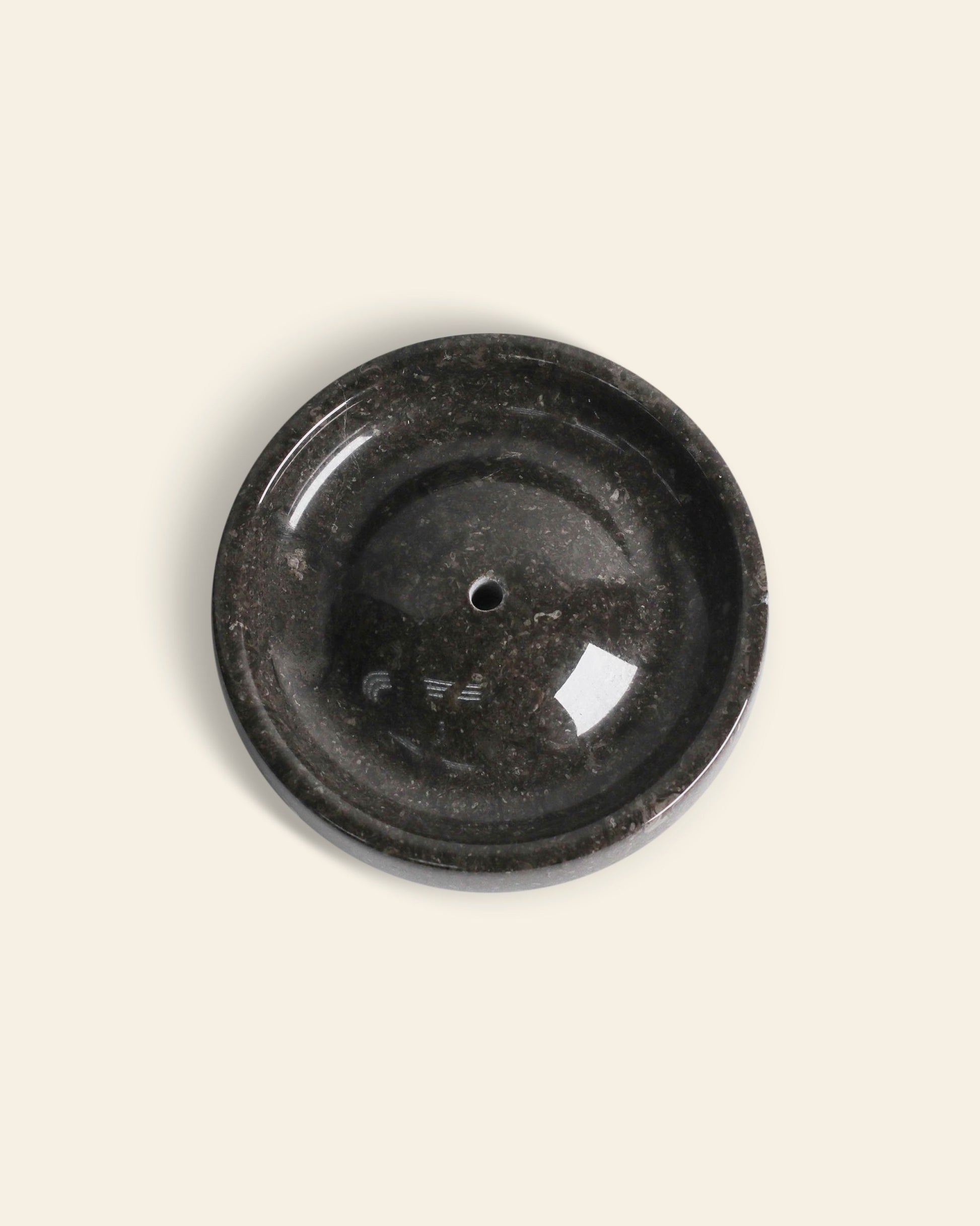 The stillgoods Ebb Incense Holder is a round, dark stone tray with a central hole, displayed from above on a light beige background.