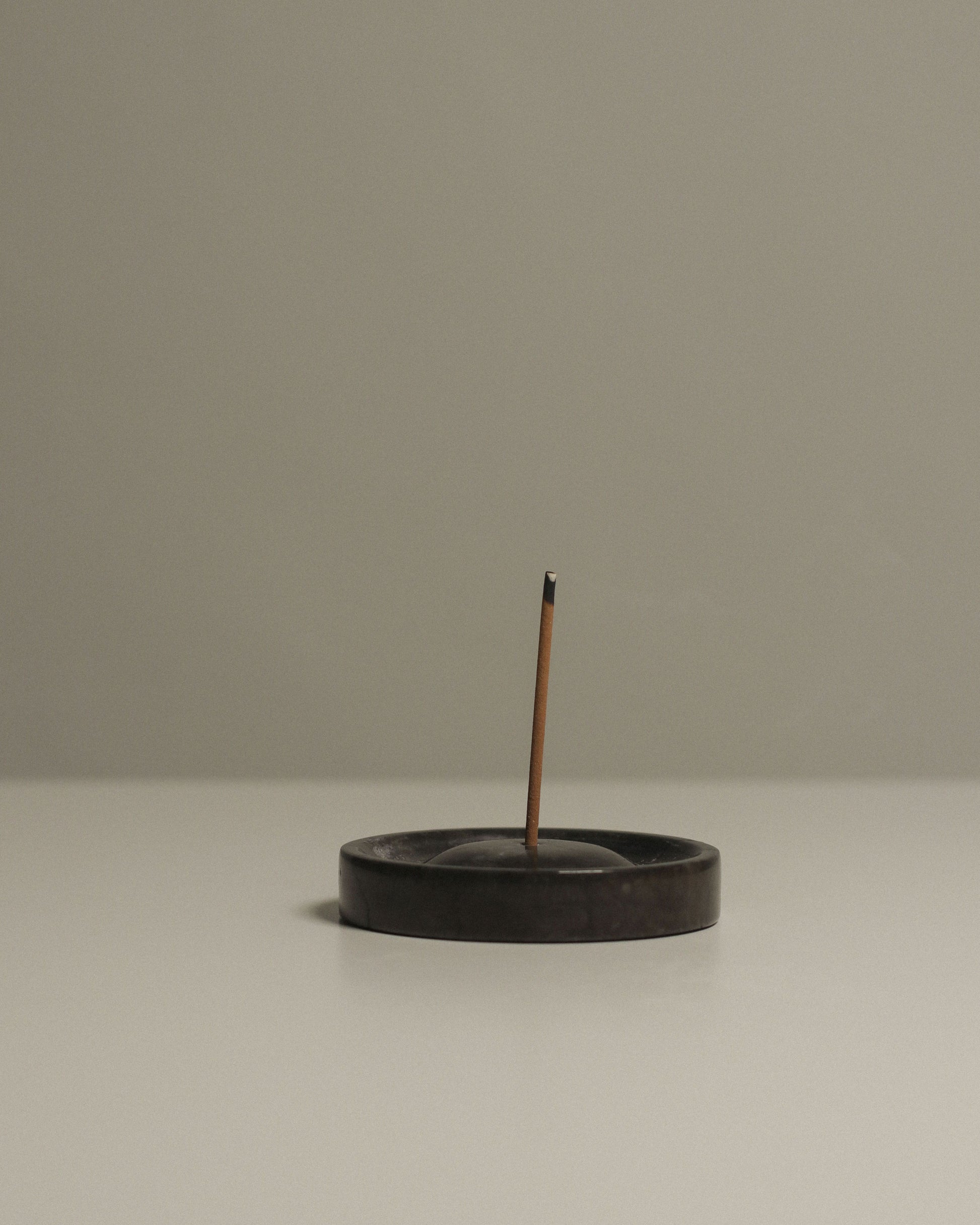 A single incense stick burns in the stillgoods Ebb Incense Holder by stillgoods, which is round and dark, resting on a smooth, light surface with a plain backdrop. Thin smoke gently rises from the tip of the incense.