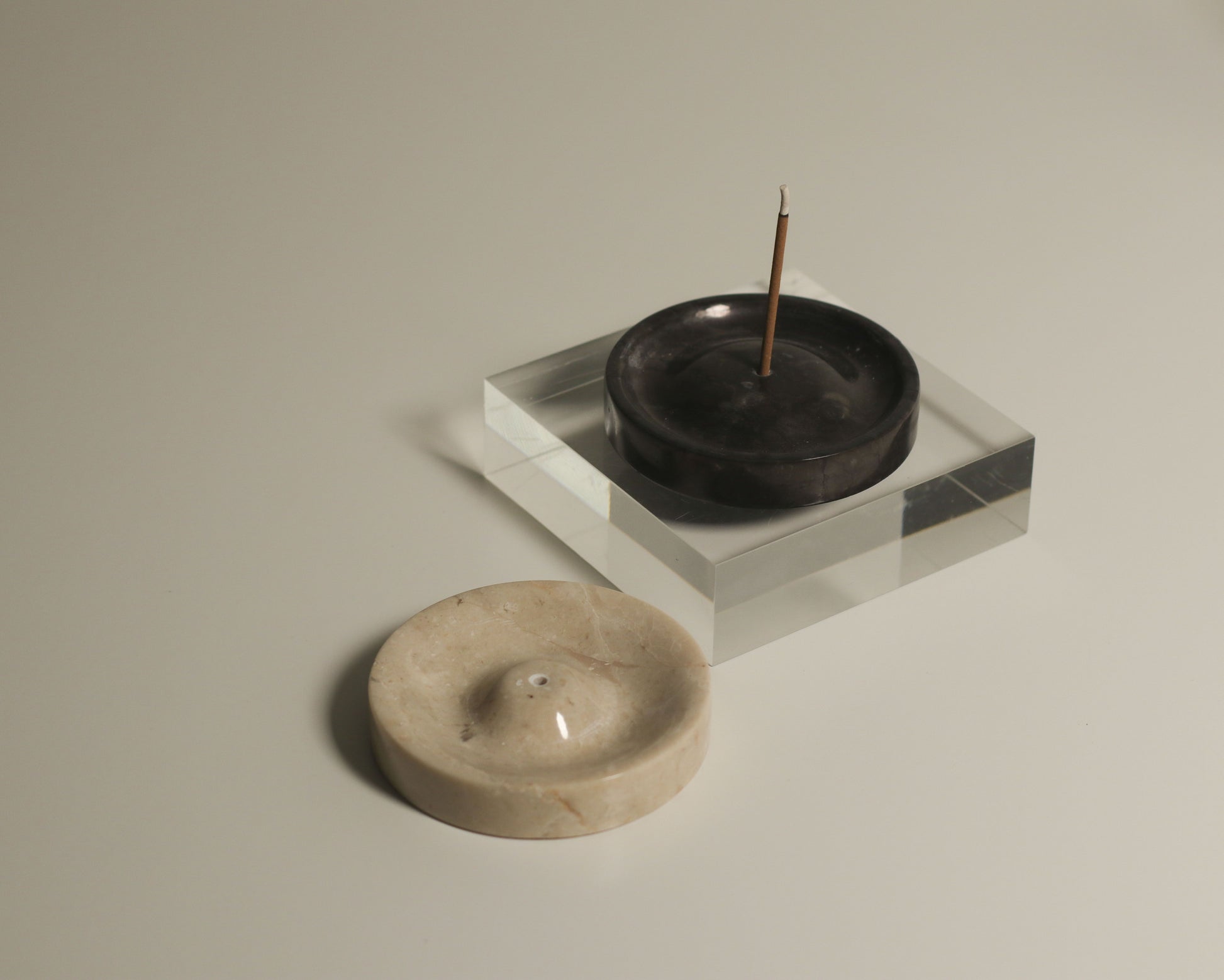 The stillgoods Ebb Incense Holder, a black round piece on a clear square base that doubles as an accessory tray, sits nearby a beige ikebana holder—both by stillgoods—adding elegant minimalist charm.